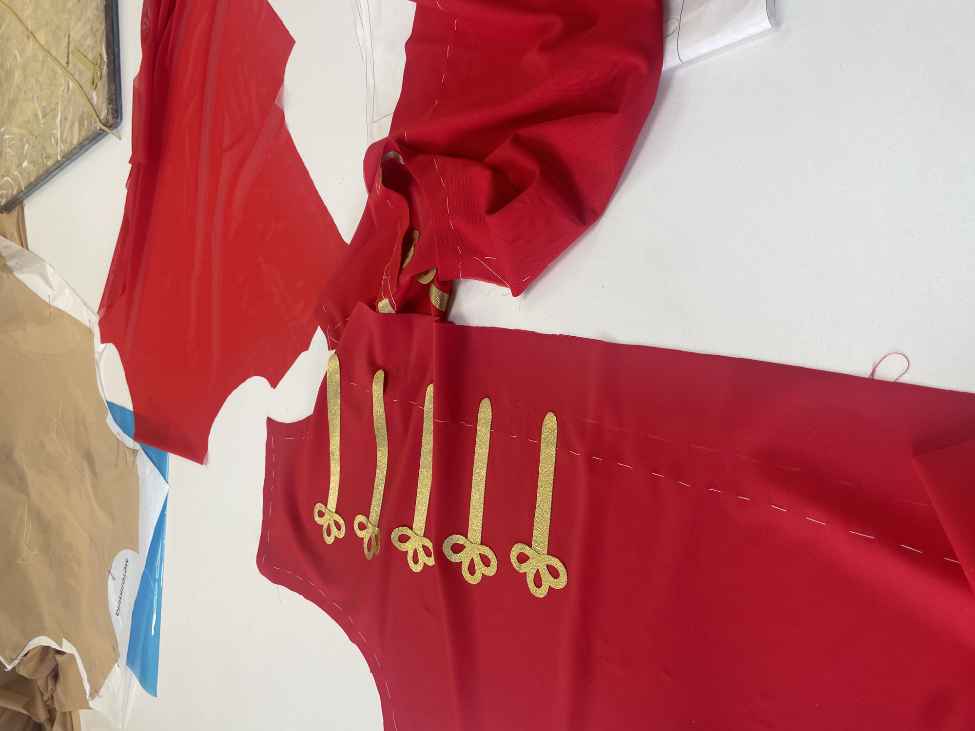 This image depicts a close-up, high-angle view of a white table covered with sewing materials, likely in a studio or workshop setting. The primary focus is on several pieces of vibrant red fabric. One large piece of fabric in the foreground shows a partial garment, possibly a jacket or coat, with five decorative gold frog closures sewn onto it in a vertical line. The fabric is laid flat and appears to be partially basted or pinned with white thread. To the right of this garment piece, another section of red fabric is draped, showing gathered seams and edges. In the background, there are pieces of sewing pattern paper, one with blue markings and text, and another in a tan color, both appearing to be draped over the table. A small, wooden block with blue fabric is visible in the upper right corner, along with a pair of gold-handled scissors resting on it. The overall scene suggests the process of creating a garment, likely a costume, given the elaborate frog closures. There are no people visible in the image. The location is not specified, and therefore cannot be confirmed to be in Saint Petersburg, Russia.