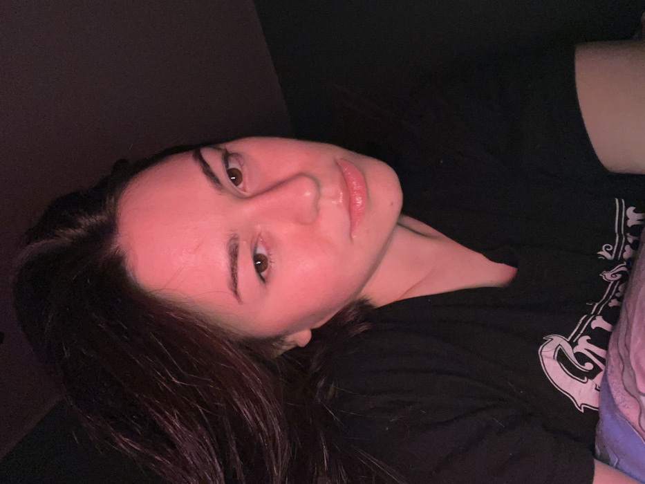 A young woman lies in bed, angled towards the camera. Her face, illuminated by a reddish light, features dark eyebrows and eyes, a prominent nose, and glossy lips. She wears a black t-shirt with a faded white design on the right side of the chest. Long, dark hair cascades around her face and shoulder. The background is mostly dark, with a glimpse of a wall behind her and a patterned blanket to her right.