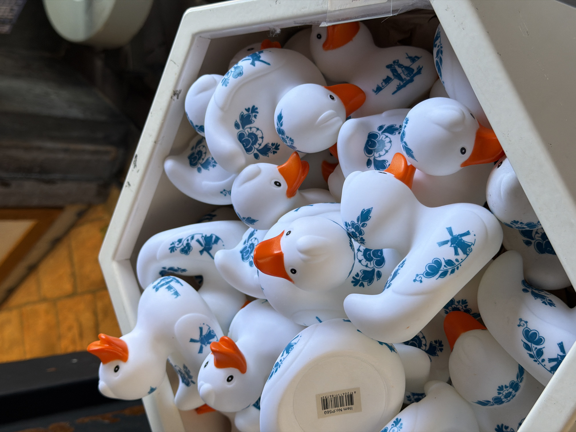 The image captures a close-up view of numerous white rubber ducks with orange beaks, densely packed within an off-white, multi-sided display container. Each duck is adorned with intricate blue patterns reminiscent of Delftware pottery, featuring various traditional Dutch motifs such as windmills, floral designs, and silhouettes of figures.
The ducks are merchandise, likely for sale or on display in a shop. There is no active event or human interaction depicted; the scene is static, showcasing the collection of identical, culturally themed toys.
Notable details include the clear, bright lighting, suggesting it is daytime. The Delftware-style decorations and the windmill motifs on the ducks strongly indicate a connection to the Netherlands, which aligns with the given location context of Amsterdam. The ducks are piled on top of each other, creating a sense of abundance.
On the underside of one duck, a white sticker is visible with a black barcode and the text "Item No: P560." The rest of the background is blurred and out of focus, showing dark, possibly wooden structures behind the container. The photograph is taken from a slightly downward angle, looking into the container.