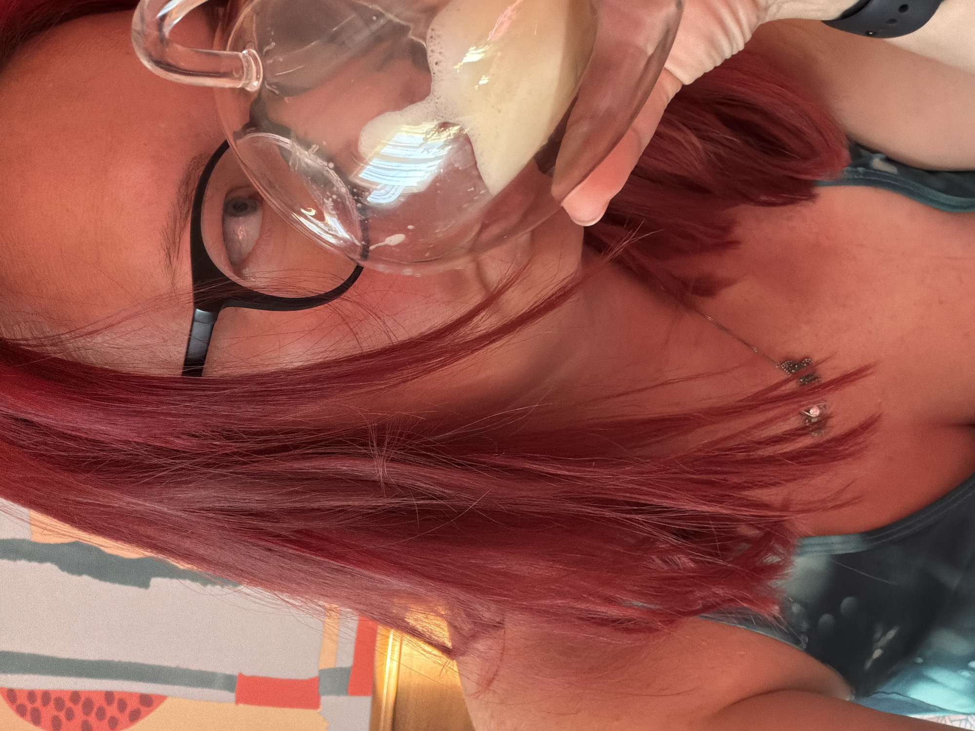 A person with red hair, wearing glasses, holds a glass containing a frothy, light-colored liquid up to their face. The person is wearing a green top. A necklace is visible on their neck. The environment appears to be indoors with a patterned wall in the background. There is no indication of the location being Chaikovsky, Russia.