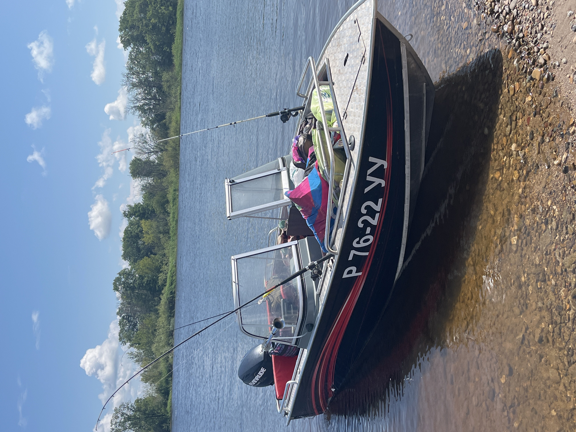 An aluminum motorboat with the registration number "P 76-22 YY" is partially beached on a stony shore of a wide, calm river under a clear blue sky with scattered white clouds. The boat, painted black with red racing stripes along the hull, has a dark grey interior with red seating visible. A black Evinrude outboard motor is mounted on the stern. Two fishing rods are set up on the boat, one on each side. Various bags and items, including a brightly colored inflatable item, are stowed in the bow of the boat. The riverbank is lined with dense green trees. The water is clear enough to see the gravelly riverbed. This scene is consistent with a location in or near Ust-Belsk, Russia, a settlement along the Angara River.