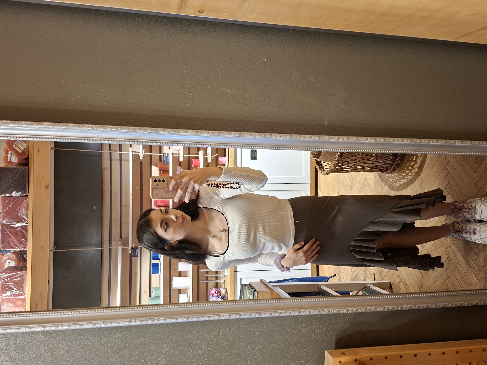A woman is taking a selfie in a mirror. She is wearing a white long-sleeved shirt and a brown skirt with ruffles. She is standing on a wooden floor in what appears to be a store. Shelves containing various items are visible in the background. The location might be a shop in Sofia, Bulgaria, given the possibility of European-style architecture or merchandise typically found in such locations.