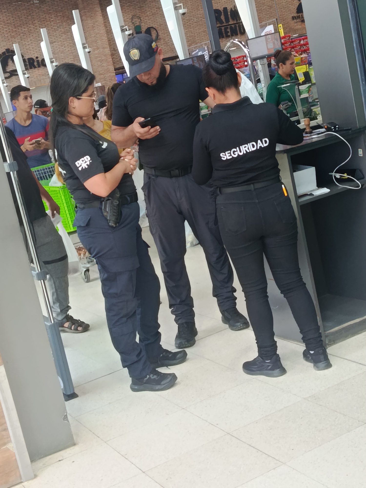 The image captures a scene inside what appears to be a supermarket or large retail store, likely at a checkout or security point. Three individuals, identified as security personnel by their black uniforms, are prominent in the foreground.
On the left, a woman wearing a black short-sleeved top with "DCD" printed on it, dark blue cargo pants, and glasses, stands with her hands clasped. A black holster with a visible firearm is strapped to her right hip. Next to her, a man in a black t-shirt, dark pants, and a dark baseball cap with an emblem (possibly depicting the Venezuelan flag colors) is looking down at his mobile phone. On the right, another woman in a black long-sleeved shirt with "SEGURIDAD" printed in white across the back and black slim-fit pants, leans over a dark counter. She appears to be interacting with something or someone at the checkout.
In the background, a young man with a green shopping basket holds a phone, and a store employee in a green polo shirt is visible behind a counter. The setting features light-colored tiled flooring, brick-patterned walls, and distinctive white, vertical structures extending upwards, possibly part of the store's architecture or signage. A sign partially visible in the background reads "RIO MENU." The overall lighting suggests it is daytime. The scene depicts a routine moment of security presence in a commercial establishment in Juan Griego, Venezuela.