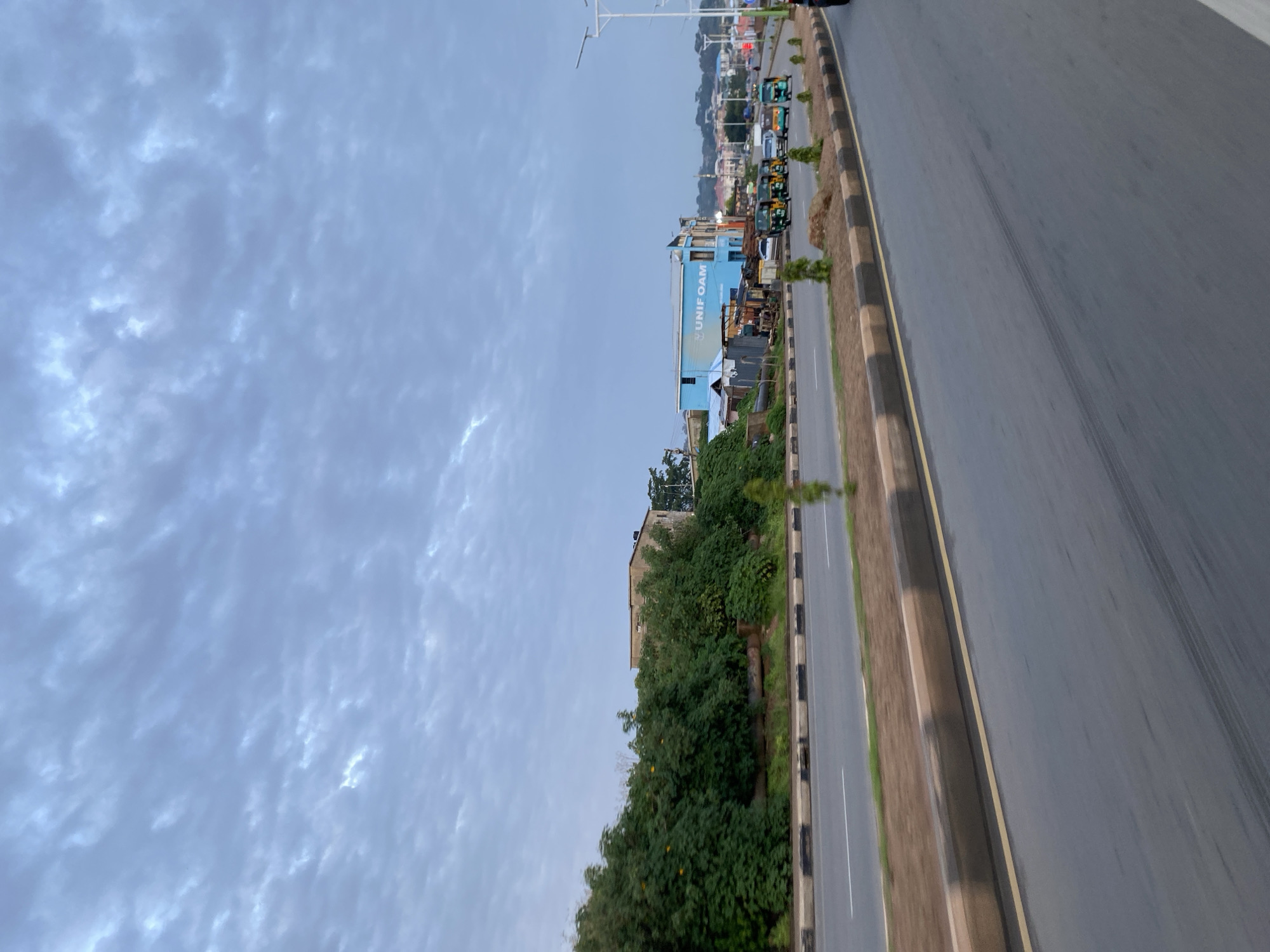 The image captures a street scene in Jos, Nigeria, under a cloudy sky. The foreground shows a two-lane road with a median strip. The road surface is wet, possibly from recent rain, and tire marks are visible. Along the median, there are small trees and decorative rocks.
On the left side of the road, a dense line of green trees and bushes forms a natural barrier. Behind the foliage, a building with a light-colored facade is partially visible.
On the right side of the road, a bright blue building with the text "UNIF OAM" is prominent. Adjacent to it are several smaller structures, some appearing to be makeshift stalls or workshops. A line of vehicles, including several auto-rickshaws (popularly known as "Keke") and cars, are traveling on the road. The background reveals a cityscape with more buildings and trees, extending towards a hilly horizon. Streetlights and traffic signals are also present, indicating an urban environment. The overall impression is that of a bustling, everyday scene in a Nigerian city.