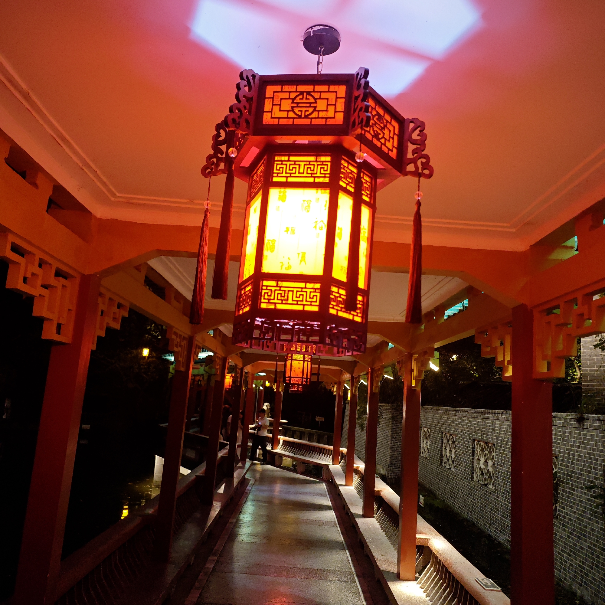 The image depicts an illuminated covered walkway in the Yuzhong District of Chongqing, China, at night. A large, ornate, rectangular Chinese lantern hangs centrally, casting a warm yellow glow. The walkway itself is framed by red wooden structures, with intricate latticework details along the top edges. Low brick walls line either side of the walkway, with occasional decorative window-like openings. The walkway appears to span a narrow body of water, which reflects some of the light. In the distance, figures can be seen on the walkway, with one person clearly visible standing and looking to the side. The overall atmosphere is warm and inviting, despite the darkness of the night.