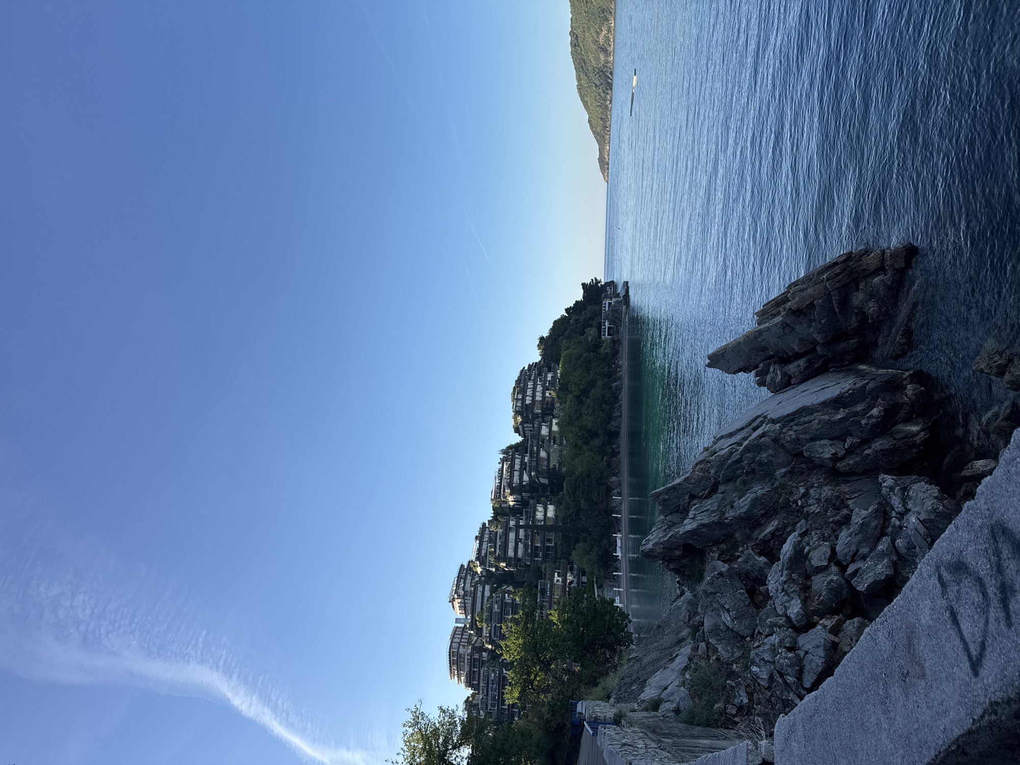 The image shows a view in Budva, Montenegro, with a focus on the Adriatic Sea and a coastal area featuring unique architecture.
**Environment:** The scene is dominated by a clear, blue sky taking up a significant portion of the frame. The Adriatic Sea is visible on the right side, its surface showing patterns of light reflecting off the water. The shoreline is rocky and rugged, with visible rock formations near the foreground. A mountainous landscape is discernible in the distance on the right, covered with vegetation.
**Subjects:** A complex of buildings is perched on the hillside to the left of the sea. The buildings are multi-story and appear to be constructed in a terraced or stepped manner, possibly apartments or a resort. The architecture seems contemporary with a dense arrangement of structures. Trees are interspersed among the buildings, adding greenery to the landscape.
**Objects:** A concrete wall or structure is visible in the lower right of the frame, displaying what appears to be graffiti. A possible boat is observed in the sea in the distance.
**Location:** The overall composition suggests the location is a coastal region known for its distinctive landscape and architectural features, consistent with the area around Budva, Montenegro.