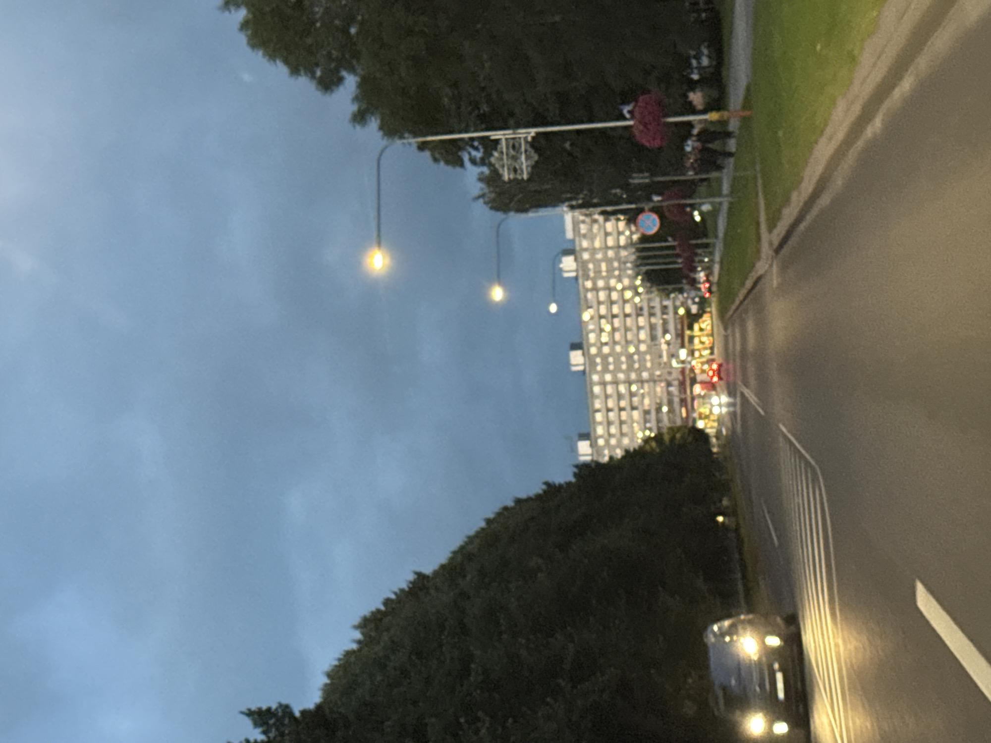 The image captures a twilight street scene in Tallinn, Estonia, focusing on a road with a car driving in the left lane. The car's headlights are illuminated, casting a yellowish glow on the asphalt and the adjacent pedestrian crossing. On the right side of the road, a sidewalk is visible, lined with streetlights and trees. Beyond the trees and sidewalk, a large, multi-story building with numerous illuminated windows dominates the background. The sky is cloudy and dark, suggesting it is either early morning or late evening. The overall impression is of a quiet urban environment under a cloudy sky.
