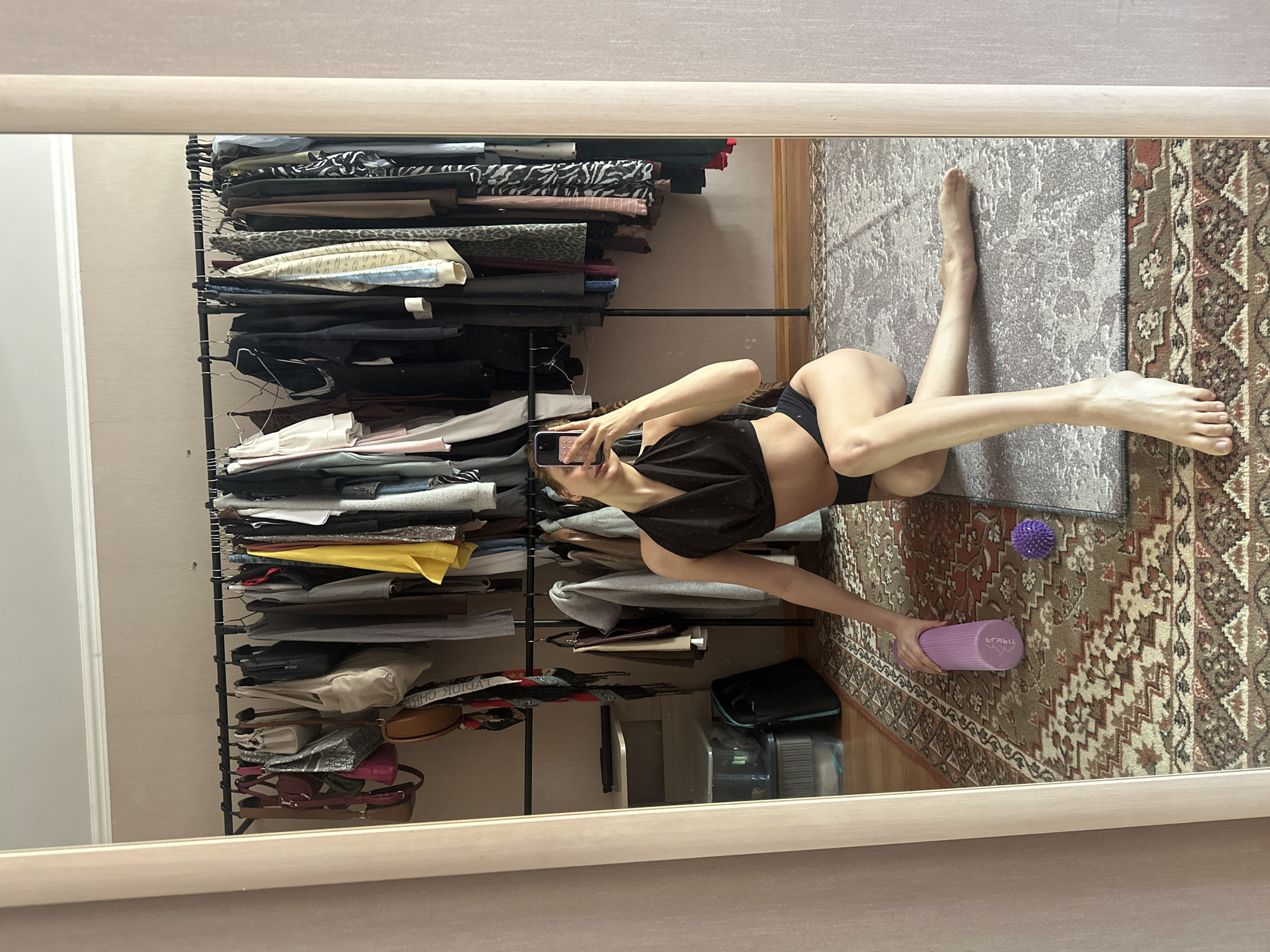 A person is reflected in a mirror. The person is wearing a black top and black shorts, holding a purple foam roller and a mobile phone. The mirror is reflecting a room with a clothing rack filled with clothes, a rug, and other items. The location appears to be indoors. There is no verifiable indication to indicate this scene is in Krasnoyarsk, Russia.