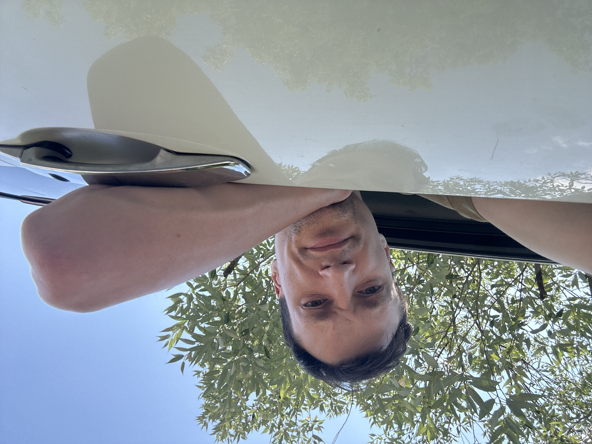 A low-angle, wide shot captures a man's face and torso as he leans over the edge of a white car with a chrome door handle. The man is looking directly at the camera with a neutral expression. Green foliage from trees and a clear blue sky are visible above him, suggesting an outdoor setting. The car's white surface reflects the surrounding environment, including trees and the sky. The image appears to be taken in Makiv, Ukraine.