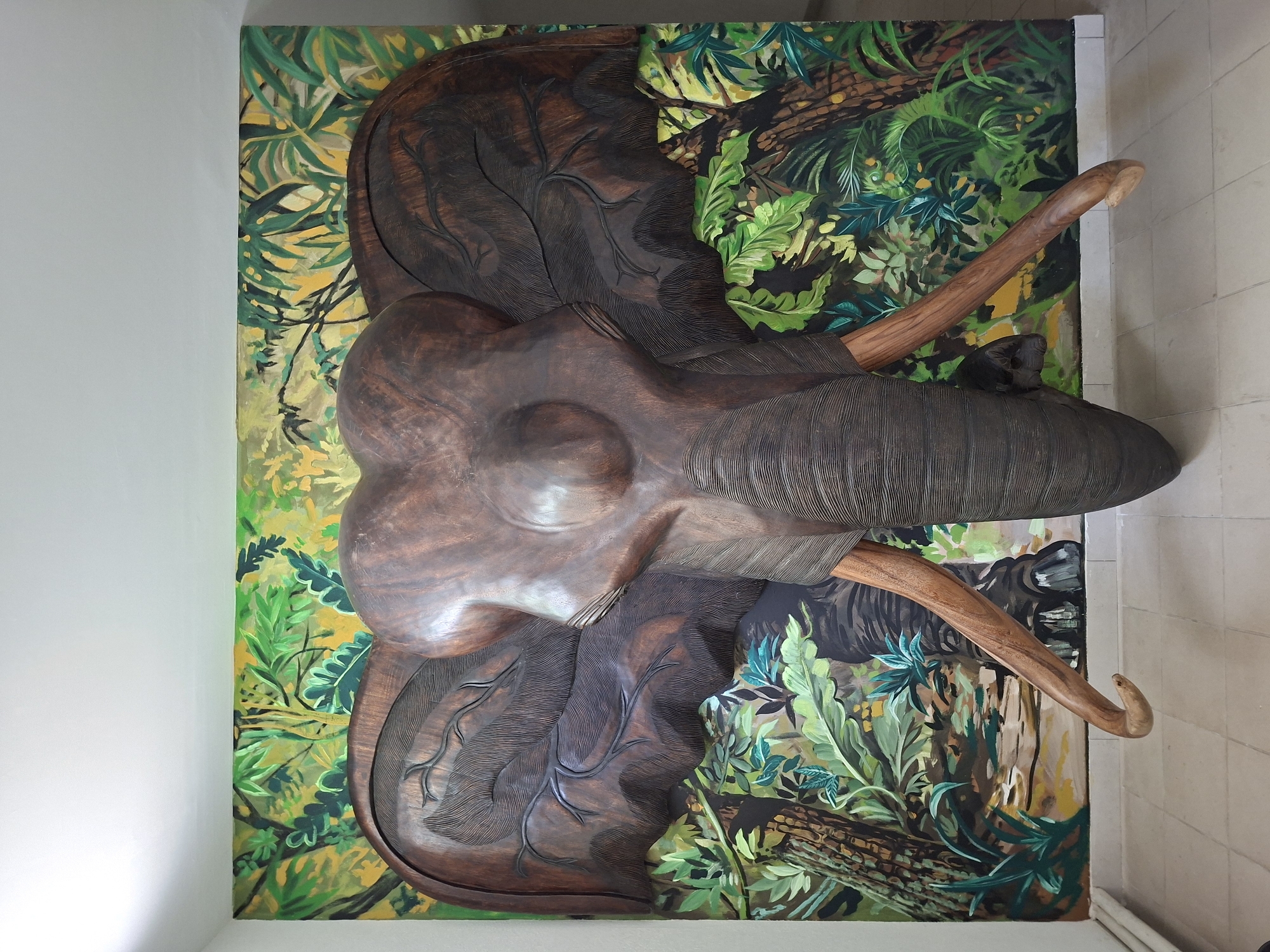 This is a medium shot, eye-level photograph of a large, carved wooden elephant head mounted on a wall painted with a jungle mural. The elephant's head is dark brown and intricately carved, with prominent tusks made of lighter wood. Its ears are spread wide, and the trunk curls downwards. The mural behind the elephant features lush green foliage and trees, with yellow and green hues dominating the background. The mural extends across the entire wall, creating a jungle scene. The elephant carving appears to be a focal point of the room. The floor is tiled with light-colored square tiles. The wall on the left is white. In the lower part of the image, a white radiator or pipe runs along the base of the wall. The overall impression is of a decorative installation, possibly in a public space or a home with an exotic theme. The location in Ialoveni, Moldova is mentioned, suggesting this might be a landmark or point of interest in that city.