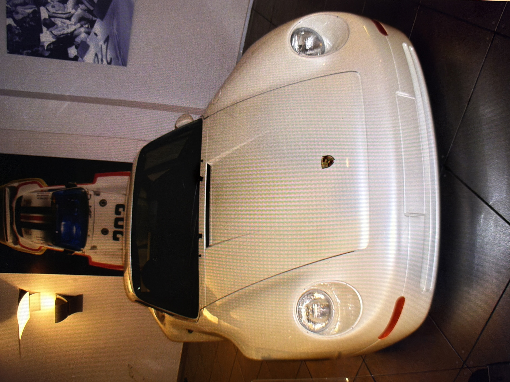 The image shows the front of a white Porsche 959. The car is parked indoors, in what appears to be a showroom or a private collection. The lighting is dim, with a warm glow from a nearby wall sconce. In the background, another car, possibly a racing car, is visible. A framed picture is hanging on the wall to the right. The floor is tiled with large, dark squares. There are no people visible in the image. The location of the image is not specified as being in Kyiv, Ukraine.