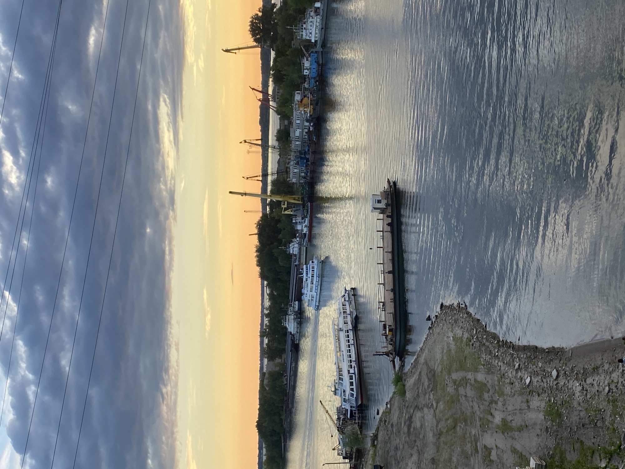 This media file shows a scenic view of a river port at sunset. The sky is a mix of dark clouds and a soft, warm orange glow, indicating the sun is low on the horizon. Several large cargo ships are docked along the riverbank, with cranes visible in the background, suggesting industrial activity. The water is calm, reflecting the colors of the sky and the shapes of the boats. On the right side of the image, a rocky embankment slopes down to the water, with some sparse vegetation. Power lines stretch across the top left of the frame, adding a modern industrial element to the natural landscape. The overall atmosphere is tranquil, with the warm hues of the sunset creating a peaceful ambiance. The scene appears to be set in a port city, likely in Russia given the provided location context. There is no visible text in the image.