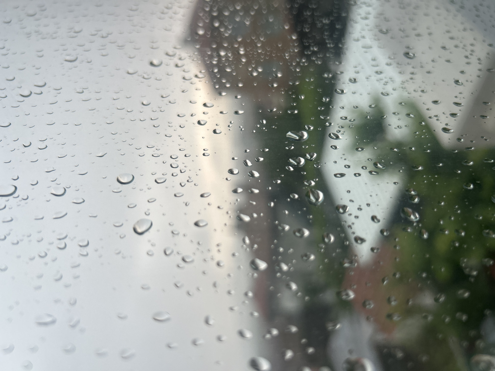 The image displays numerous raindrops of varying sizes adhering to a transparent surface, most likely a window or windshield. The water droplets in the foreground are in sharp focus, showcasing their individual shapes and reflections of the ambient light.
Behind the rain-covered surface, the background is significantly blurred, indicating a shallow depth of field. Indistinct shapes on the left and center suggest structures or buildings with muted grey and brownish tones. On the right side, blurred patches of green foliage, likely trees or bushes, are visible. The overall lighting suggests an overcast sky during daytime.
The scene clearly depicts rainy weather conditions. The perspective is from an interior looking outwards, creating a contemplative mood through the obscured view. There is no visible text in the image. This scene is set in Aytos, Bulgaria.