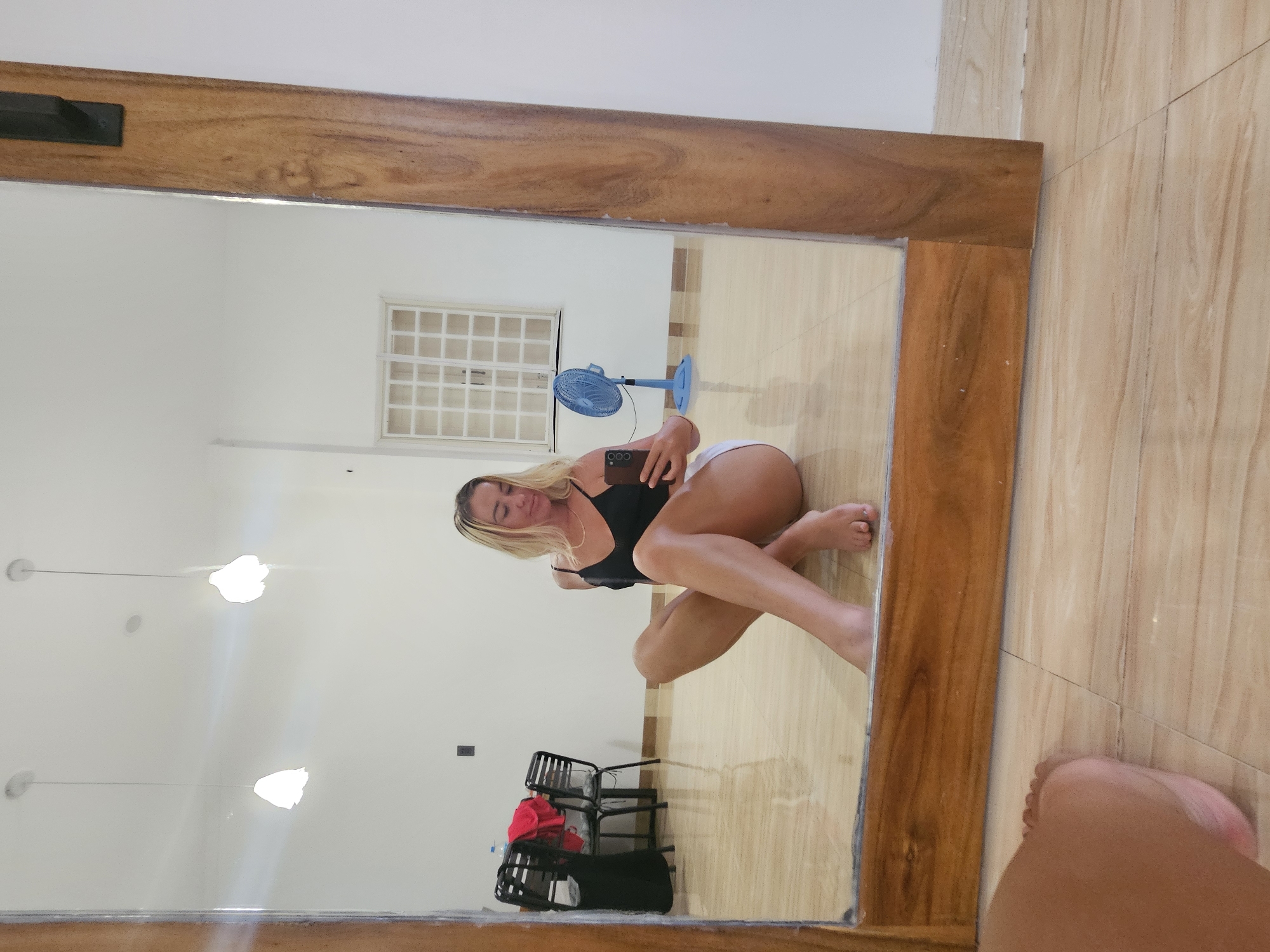 A young woman with blonde hair is sitting on the floor in a room, holding a smartphone and taking a selfie in a large, wooden-framed mirror. She is wearing a black tank top and white shorts. The room has a light-colored tiled floor and white walls. In the reflection of the mirror, there are two pendant lights hanging from the ceiling, a window with a grid pattern, and a blue fan. In the background, there are some chairs with clothes draped over them and a water bottle. The image appears to be taken in a residential or studio space within Phu Thuy Ward, Vietnam.