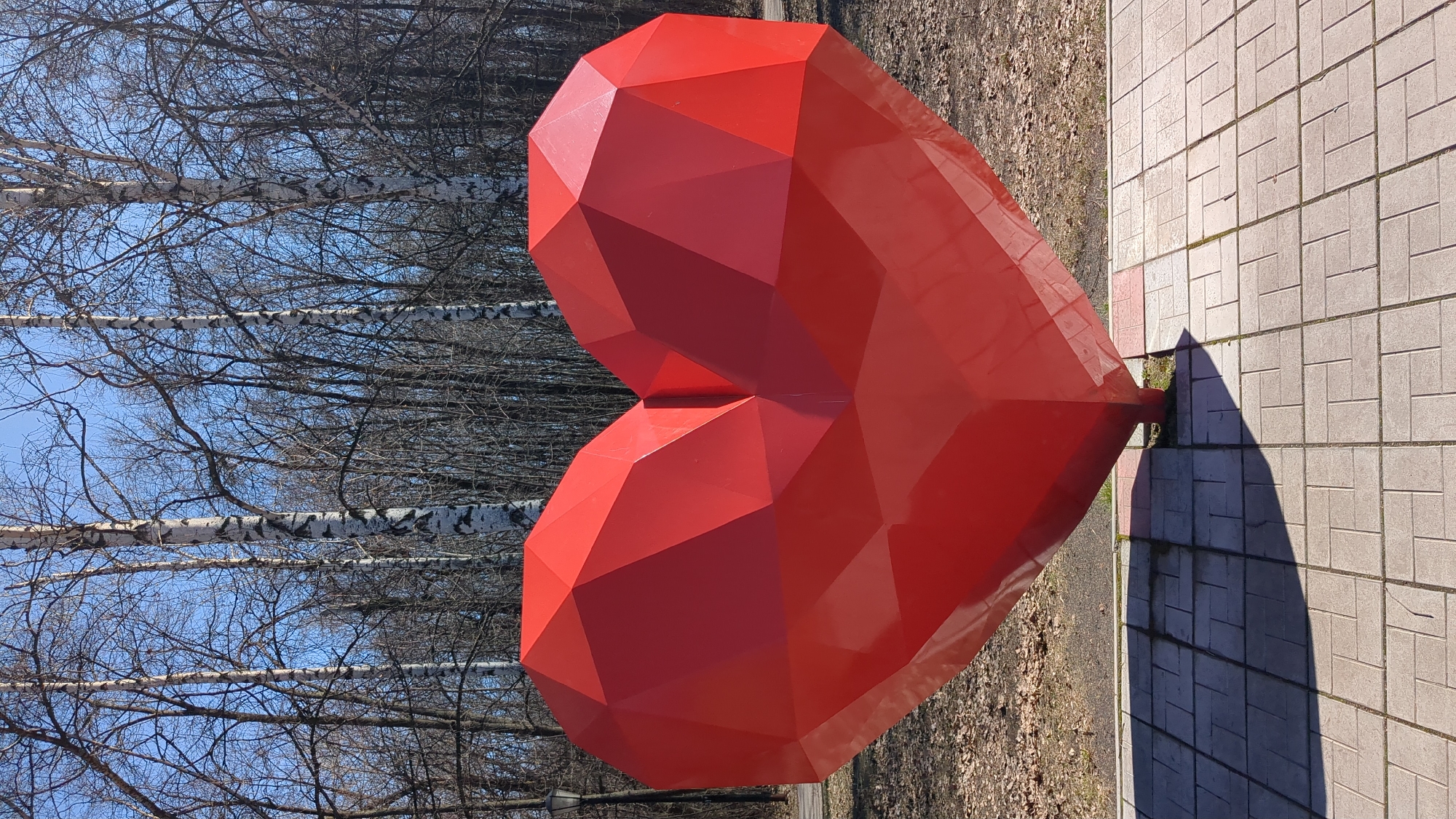 A large, red, faceted heart sculpture is positioned on a paved square in the city of Ulyanovsk, Russia. The sculpture casts a dark shadow on the light grey paving stones. In the background are leafless birch trees against a clear blue sky. The area appears to be a park or public space.