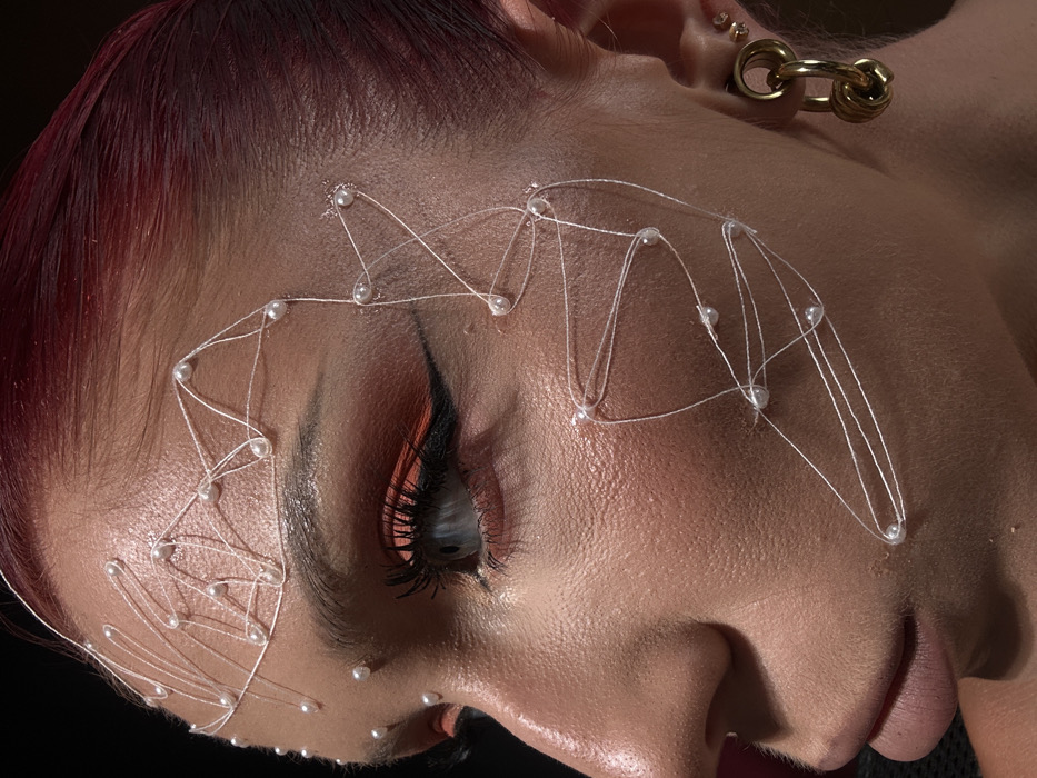 A close-up profile view captures the right side of an individual's face in the city of Dalung, Indonesia. The person has red hair, a blue eye, and full lips. The skin exhibits a matte finish with visible pores and slight sheen on the cheekbone. Intricate facial adornment consists of white thread intricately arranged across the forehead, temple, and right cheek, secured by multiple small, spherical white beads, resembling pearls. Eye makeup includes a bright orange eyeshadow and a sharp black winged eyeliner, complementing prominent dark eyelashes. The right earlobe features a gold-toned ear gauge or tunnel, from which a large gold-toned earring composed of two interlocked thick rings dangles. Two smaller stud earrings are visible higher on the ear. The background is uniformly dark, providing sharp contrast to the illuminated subject.