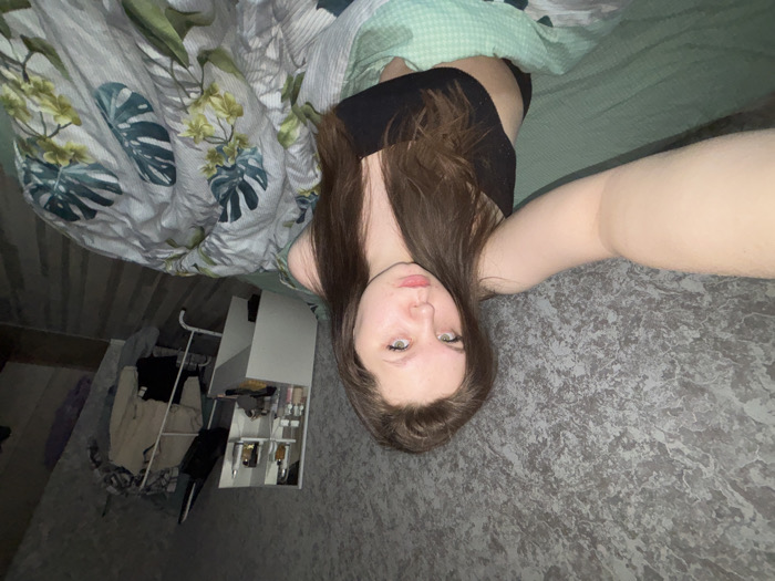 The image depicts an indoor scene, likely a residential bedroom, within an unspecified location in Perm, Russia. A young adult female, with long brown hair, light-colored eyes, and fair skin, is the central subject. She is wearing a black strapless top or bra and is positioned on a bed, partially covered by bedding. Her right arm is extended, holding a device to capture a selfie, and she is looking directly into the camera with a neutral expression.
The bed is covered with a light green sheet and a duvet featuring a white background with a prominent tropical leaf pattern in shades of green and teal. To the left of the subject, a wall is visible, covered in grey wallpaper with a mottled, textured appearance resembling stone or concrete. To the right of the subject, in the background, a white multi-tiered shelving unit or vanity table holds various small bottles and containers. Adjacent to this, a white metal clothes rack is present, draped with several items of clothing, including a light-colored top and a dark lower garment. The floor in this area is a light-colored wood laminate. The overall lighting appears dim, suggesting ambient light sources.