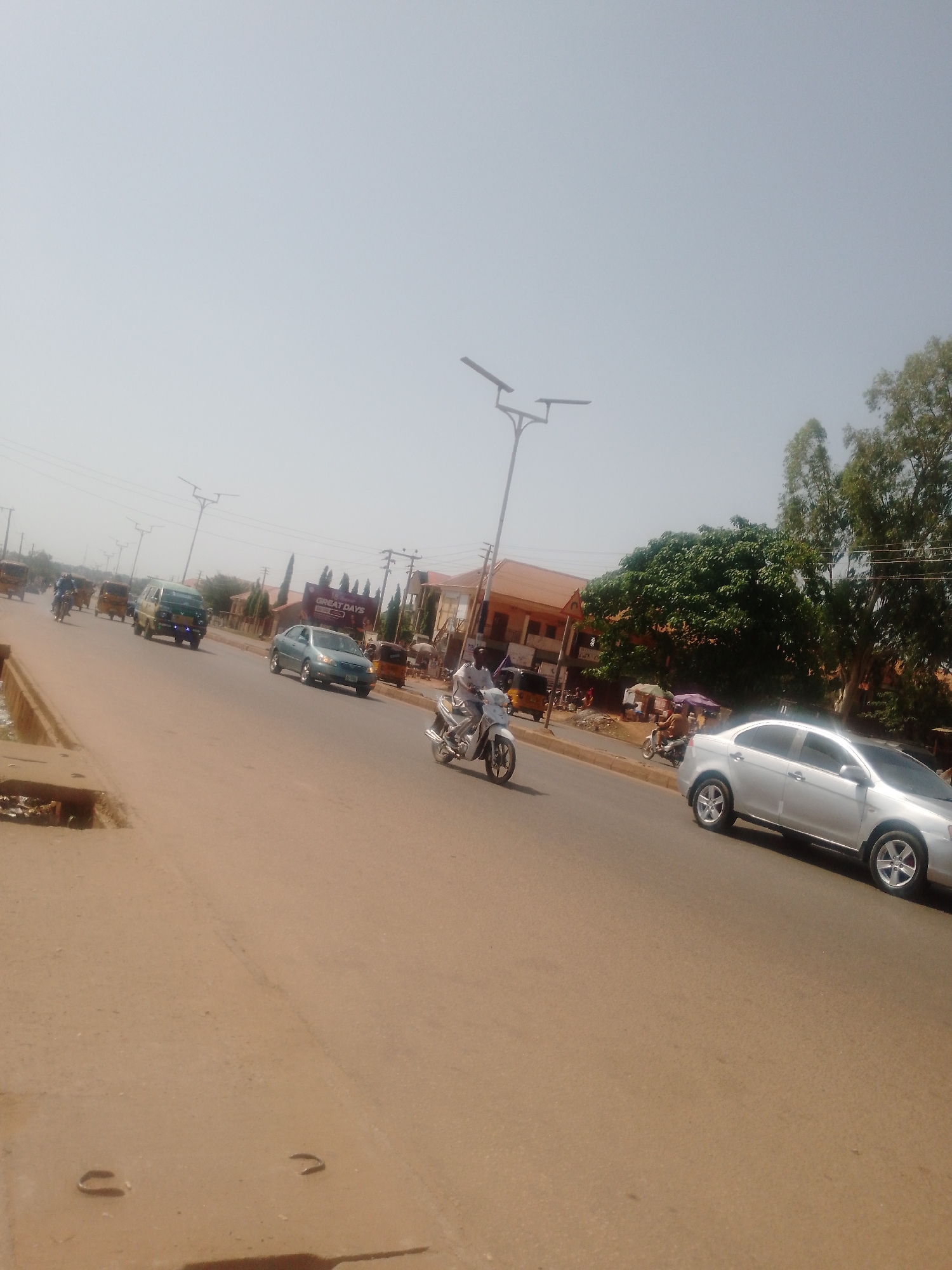 The image depicts a street scene in Sabon Tasha, Nigeria. The road is paved and appears to be well-traveled. Several vehicles are visible, including cars, a van, and motorized tricycles (keke). A person is riding a motorcycle. Buildings and trees line the street. The sky is bright and hazy, suggesting it is daytime. There is street lighting. The presence of commercial activity suggests a mixed residential/commercial area.