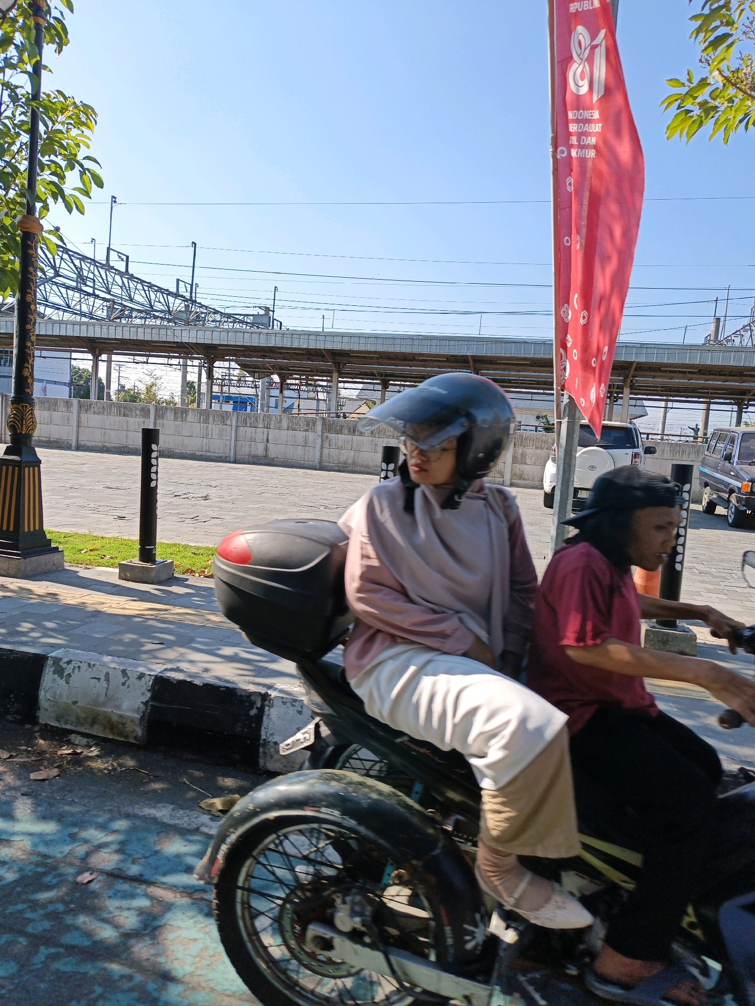 Two individuals are riding on a motorcycle in Klaten, Indonesia. The person in front, wearing a pink hijab and white pants, is driving the motorcycle. They are wearing a black helmet with a clear visor and glasses. The person in the back, wearing a maroon t-shirt and black pants, is looking to their right.
The motorcycle has a black storage box on the back. They are on a paved road with a sidewalk on the left. In the background, there are buildings, a fence, a large metallic structure that appears to be part of a transportation hub or factory, and trees. A red flag with white writing is hanging from a pole on the right side of the image. The sky is clear and blue. The lighting suggests it is daytime, with the sun casting shadows on the ground.