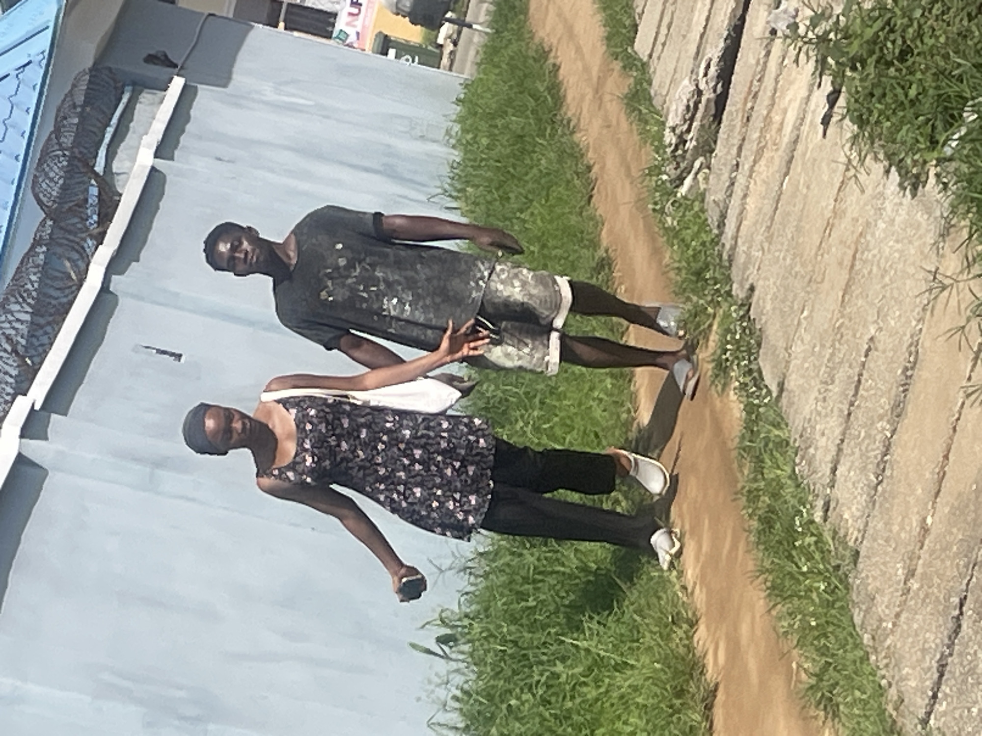 Two individuals, a male and a female, walk side-by-side along a dirt path next to a grassy area. The female is on the left, wearing a dark floral print dress, black pants, and white slip-on shoes. She is holding a dark object in her right hand and has a white bag slung over her left shoulder. The male is on the right, wearing a dark, speckled t-shirt, camouflage-patterned shorts, and silver slip-on sandals. His hands are clasped together in front of him. They are walking away from the camera, towards a light blue wall with barbed wire along the top. The environment appears to be an urban or suburban setting in Abuja, Nigeria, characterized by the wall, vegetation, and paved walkway in the foreground. The lighting suggests it is daytime, with shadows cast on the ground.