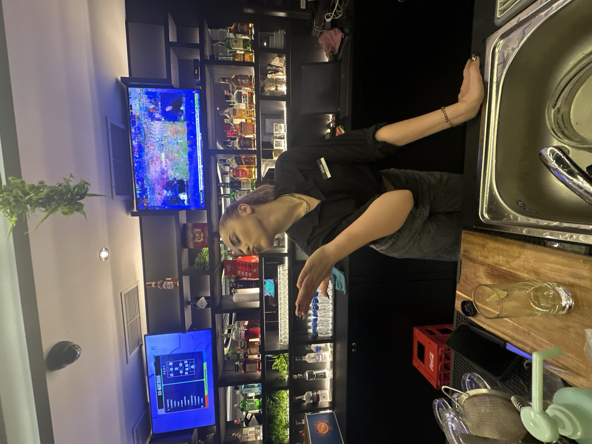 This is a vertically oriented, eye-level medium shot of a young woman working behind a bar. She is wearing a black shirt and grey jeans, with a name tag that reads "efbet". Her hair is dyed purple and she is wearing a silver chain necklace. She is leaning forward, looking down at a bottle of water on the shelf, and her right arm is extended, about to pick it up. Her left hand is resting on the edge of a stainless steel sink.
The bar is well-stocked with various bottles of alcohol on shelves behind her. A large television screen displays a soccer game, specifically showing the FIFA World Cup 2020 with Russian commentary. There is also a smaller screen displaying what appears to be a menu or game interface. In the foreground, there's a wooden countertop with a glass of liquid, a red Coca-Cola crate, and bar tools. The lighting appears to be artificial and dim, suggesting it is nighttime or indoors. The overall scene depicts a bartender actively working in a bar environment.
