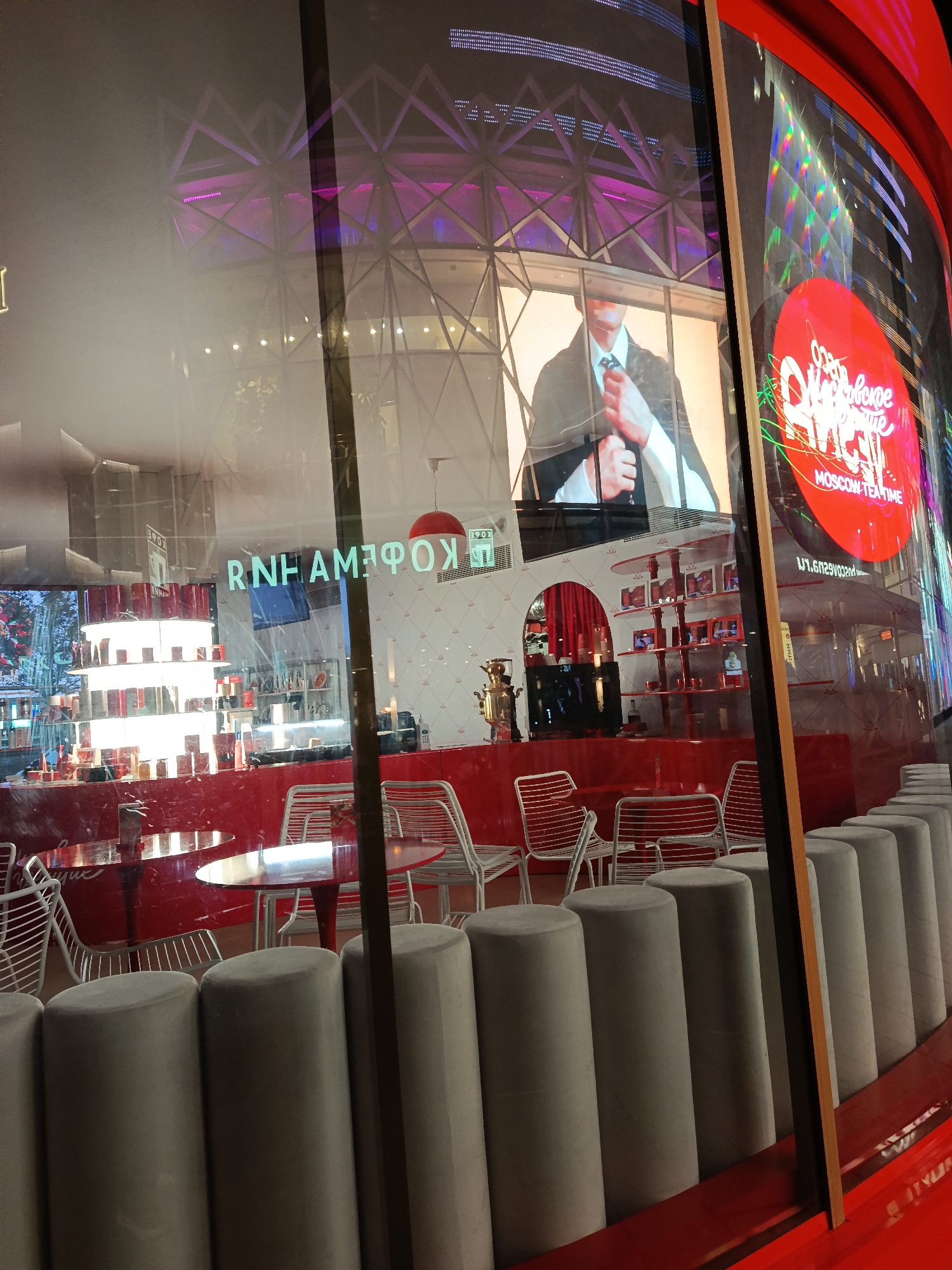 The image is a view through the glass storefront of a cafe, reflecting a building exterior with digital screens. The cafe's interior features red counters and tables with white chairs, along with cylindrical gray bar stools. A sign that reads "RNHAMЗФОK" is visible on the glass. A display shelf is filled with merchandise. The reflection shows an advertisement and another structure, with purple lighting. A red sign reading "MOSCOW TEATIME" is also reflected in the glass. Based on this, the image is likely taken in a modern, commercial area of Moscow, Russia.