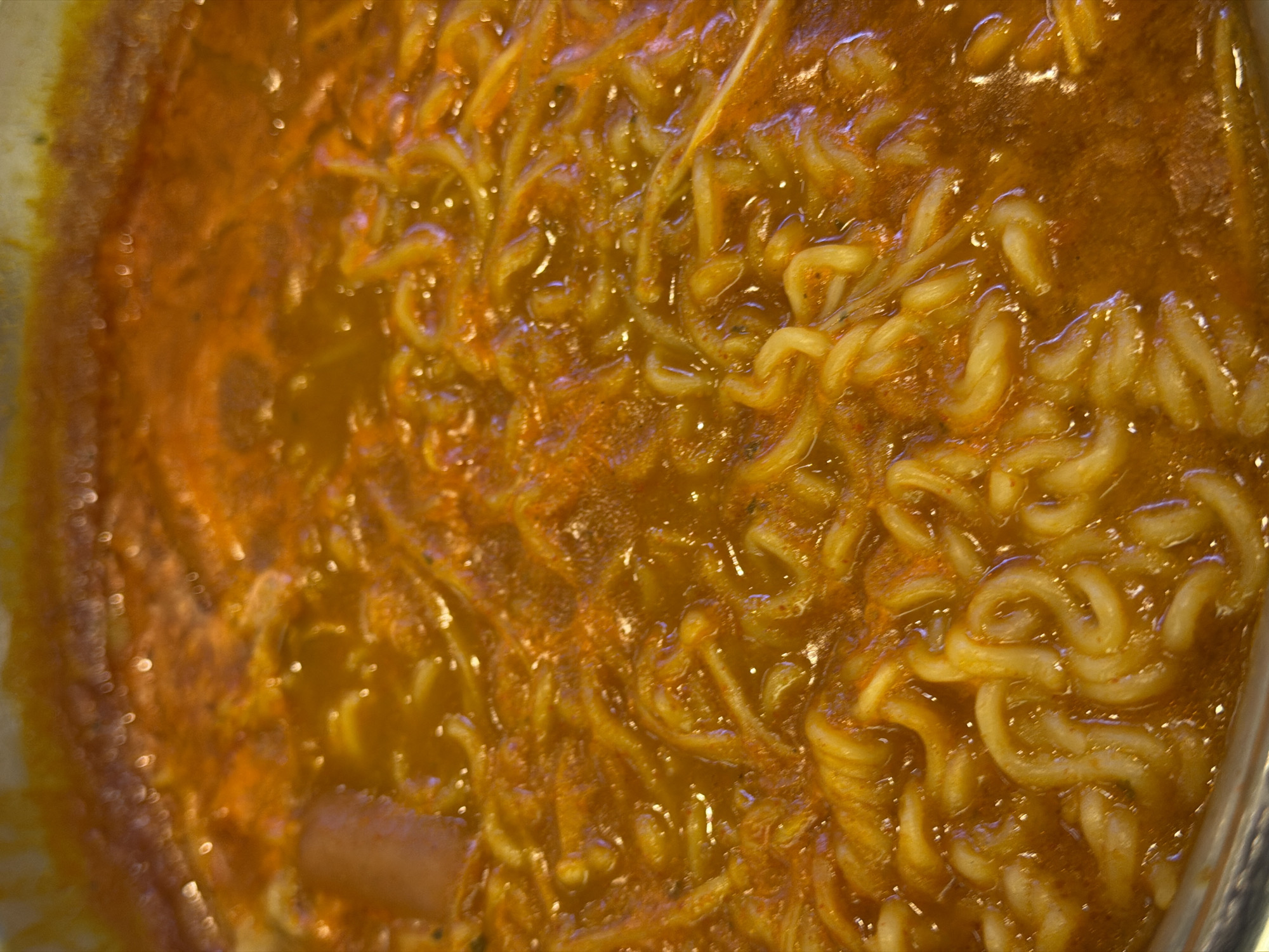 A close-up, overhead shot captures a pot of ramen noodles simmering in a rich, orange broth. The noodles, a bright yellow, are tightly coiled and submerged in the liquid, with some strands peeking above the surface. The broth appears to be seasoned with red spices, creating a slightly textured, opaque appearance. The lighting in the image creates small, bright reflections on the surface of the broth. The scene is devoid of any human presence or identifiable landmarks, making it impossible to determine its specific location within Đà Nẵng, Vietnam, or any other city.