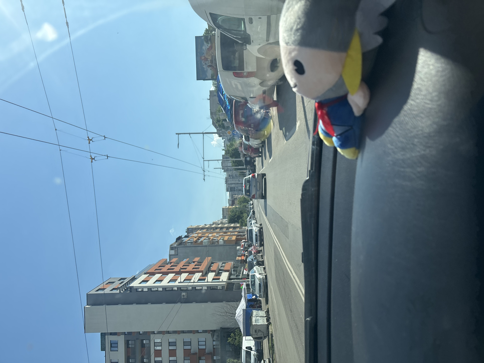 A POV shot from inside a car looking forward, the car is in traffic on a sunny day. The scene is a street in Sofia, Bulgaria, with buildings lining both sides of the road. The buildings are a mix of modern and older architecture, with several multi-story apartment buildings visible. There are cars, vans, and a bus on the road, suggesting moderate to heavy traffic. Overhead wires for public transportation, likely trolleybuses or trams, are strung across the street. In the foreground, on the dashboard, a stuffed toy resembling a superhero with a red cape, blue suit, and yellow accents is visible. The sky is clear and blue, with some lens flare from the sun.