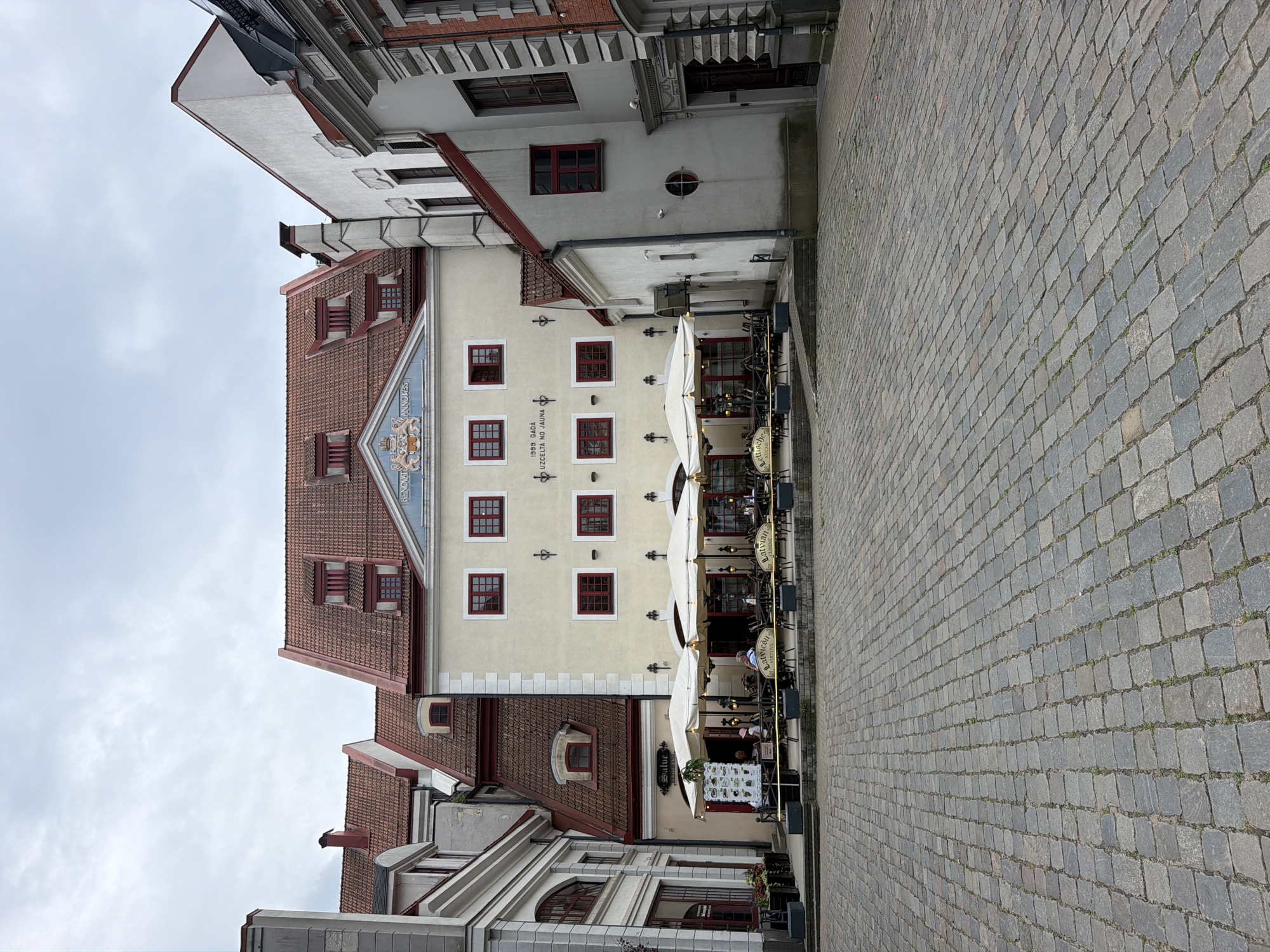 The image depicts a cobblestone plaza in the Old Town of Riga, Latvia. In the background, there are several historic buildings with red-tiled roofs and cream-colored facades. One building features a crest and the inscription "1999. GADA UZCELTA NO JAUNA". An outdoor seating area for a restaurant, with tables, chairs, and white umbrellas, is set up in front of one of the buildings. A few patrons are seated at the tables. The sky is overcast.