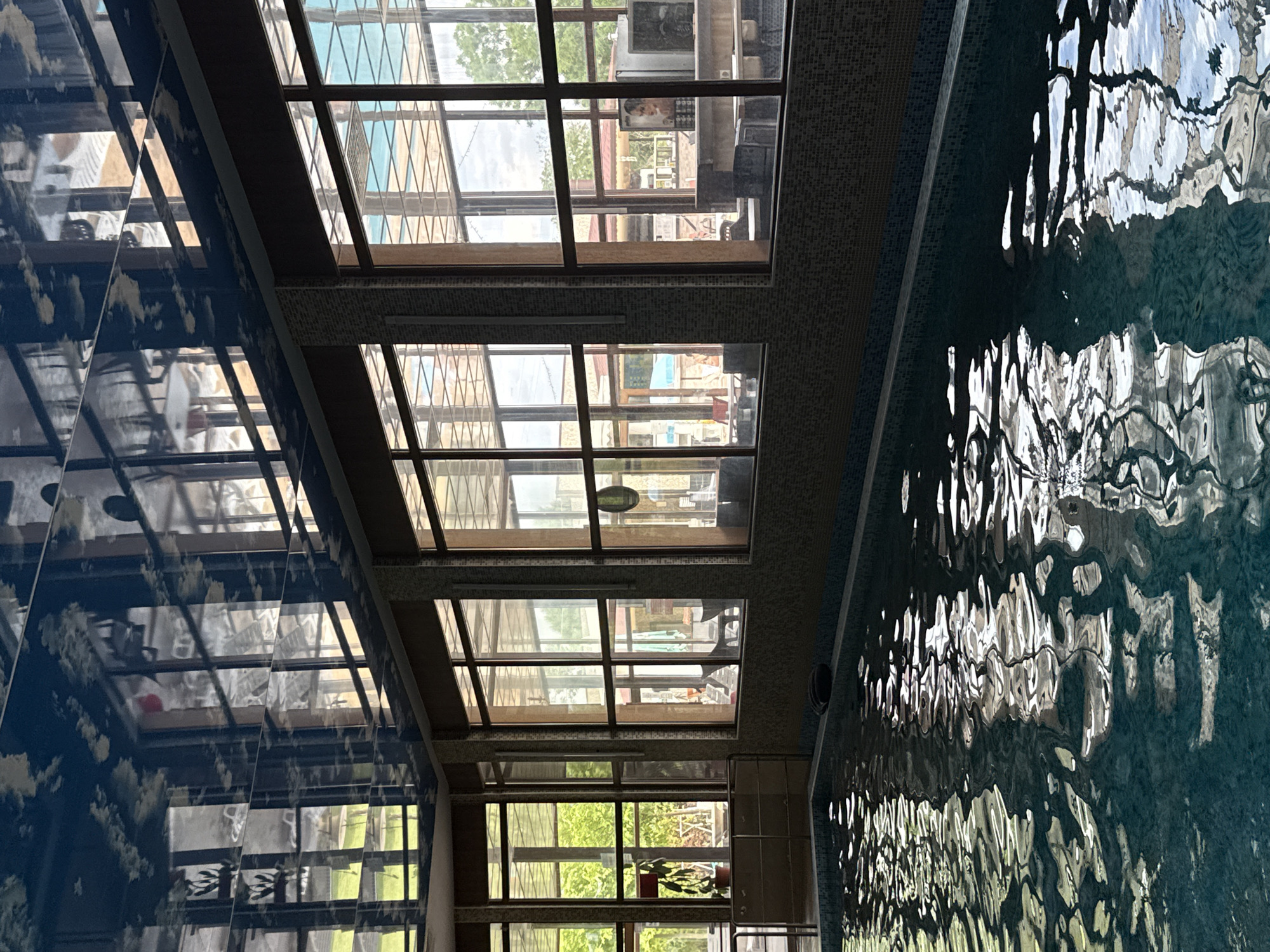 This image depicts an indoor swimming pool facility located in Tulovo, Bulgaria. The foreground is dominated by a rectangular swimming pool filled with light blue-green water, its surface actively rippling and reflecting the overhead structures and outdoor light. The pool's perimeter features a dark tiled edge, with a lighter blue strip directly below it, and a dark, possibly inflatable object resting on the edge in the middle-right. A metallic handrail is present on the left side of the pool.
A wall constructed of small, variegated brown and beige square tiles forms the background to the pool, extending upwards to a series of large, dark-framed windows. These windows illuminate the space with natural light and reveal an outdoor setting. Through the windows, lush green trees and foliage are visible, particularly on the left and right. Outdoor seating areas, comprising tables and chairs, are discernible, with faint human figures appearing to be seated at some tables in the mid-ground. A potted plant with green leaves stands to the left of the leftmost window. To the right, an outdoor counter or bar area is visible, featuring a monitor displaying a human face, and dark furniture or storage boxes beneath it. A red-roofed structure and a blue canopy are also visible through different window panes. A circular object, possibly a wall clock, is reflected on the ceiling and also visible through the windows on an interior wall beyond the outdoor area.
The ceiling is highly reflective, appearing dark blue with lighter, abstract patterns resembling clouds, and acts as a mirror, reflecting the window grid, the outdoor environment, and various indoor elements in an inverted and distorted fashion. The indoor area receives significant natural illumination from the exterior, creating a contrast with the relatively shadowed interior.