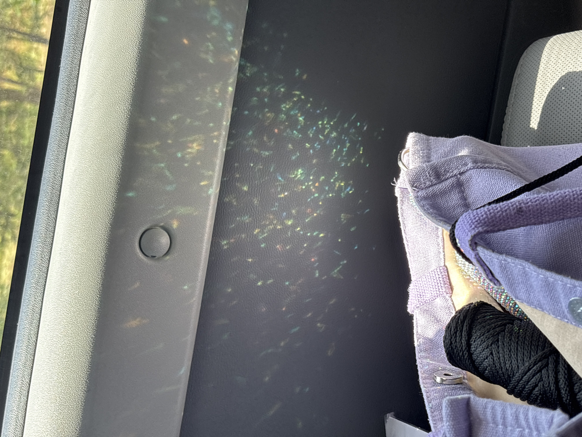 The image displays a close-up view of a car's interior, focusing on a light-colored, textured dashboard and a portion of a purple canvas tote bag. Scattered across the dashboard are numerous small, shimmering reflections of light, appearing as colorful speckles of green, blue, yellow, and red. A circular indent, possibly a button or a small compartment, is visible on the dashboard. The tote bag, partially visible in the foreground, contains a tightly wound spool of black cord. The background, visible through the car window, shows blurred greenery, suggesting an outdoor environment. The overall scene captures the effect of sunlight interacting with the car's interior surfaces. The location is not specified in the image.