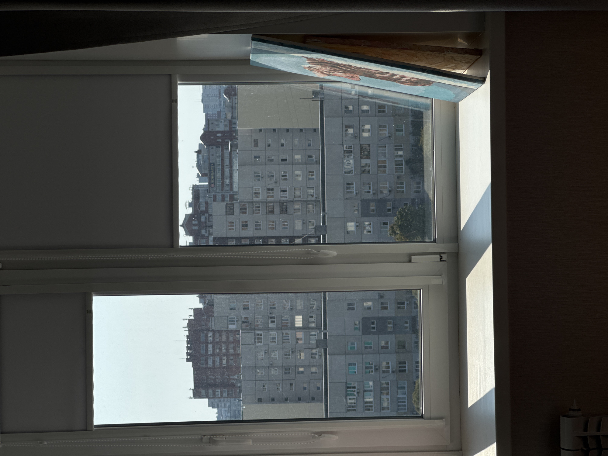 A medium shot captures a view from a window, looking out onto a city skyline. The window is divided into two panes by a white frame, with roller blinds partially covering the top of each. In the left pane, a cluster of tall, multi-story buildings with red-brown facades are visible against a pale blue sky. The right pane shows more uniform, gray apartment buildings stretching across the horizon, some with visible balconies and windows. A painting, propped up on the windowsill, leans against the window frame on the right side. The painting appears to be an abstract depiction with a dominant blue background and hints of orange and brown. Sunlight streams in from the left, casting a strong diagonal shadow across the white windowsill. The windowsill itself is clean and uncluttered, save for the painting. A radiator is partially visible in the lower left corner of the frame, adding a domestic element to the urban landscape. The overall impression is one of a quiet moment, observing the city from a comfortable indoor vantage point.