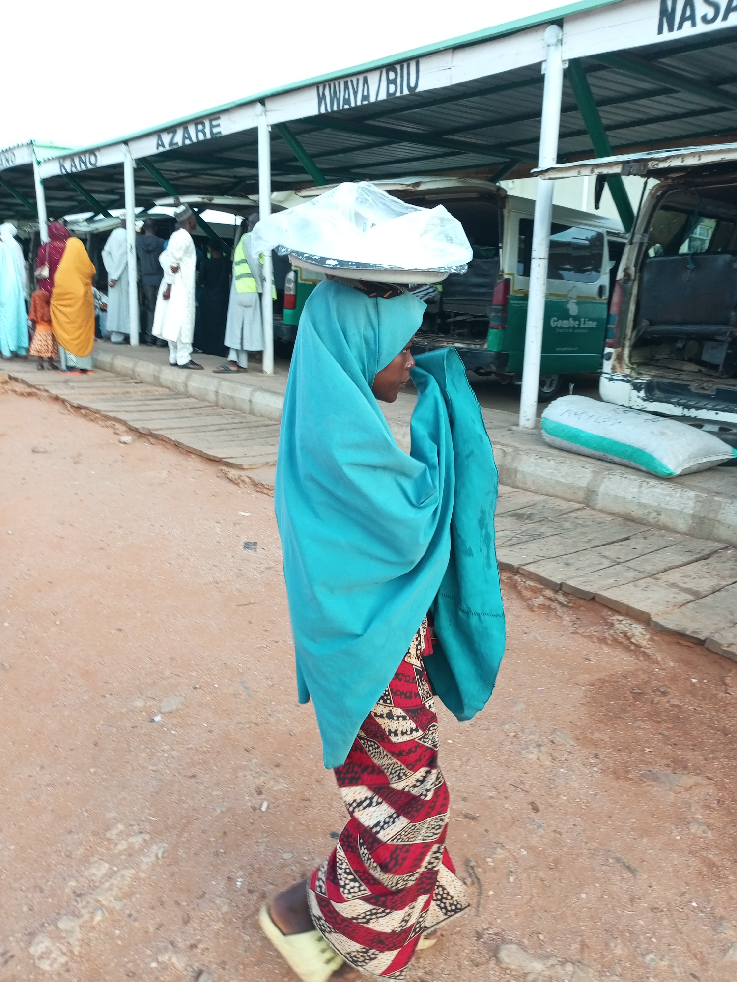 The image shows a woman in a blue head covering and a red, white, and black patterned skirt walking on a dirt road next to a bus station. The bus station has a corrugated metal roof supported by white posts, with signs indicating locations such as "Kano," "Azare," "Kwaya/Biu," and "Nasarawa." Several people are standing or sitting under the shelter of the bus station, some near parked vehicles. One vehicle in the background has "Gombe Line" written on its side. Bags of goods are visible on the ground near the bus station. The woman is carrying a large, flat object covered in plastic sheeting on her head. Her expression is neutral, and she is looking to her left. The overall scene suggests a bustling transportation hub in Gombe, Nigeria.