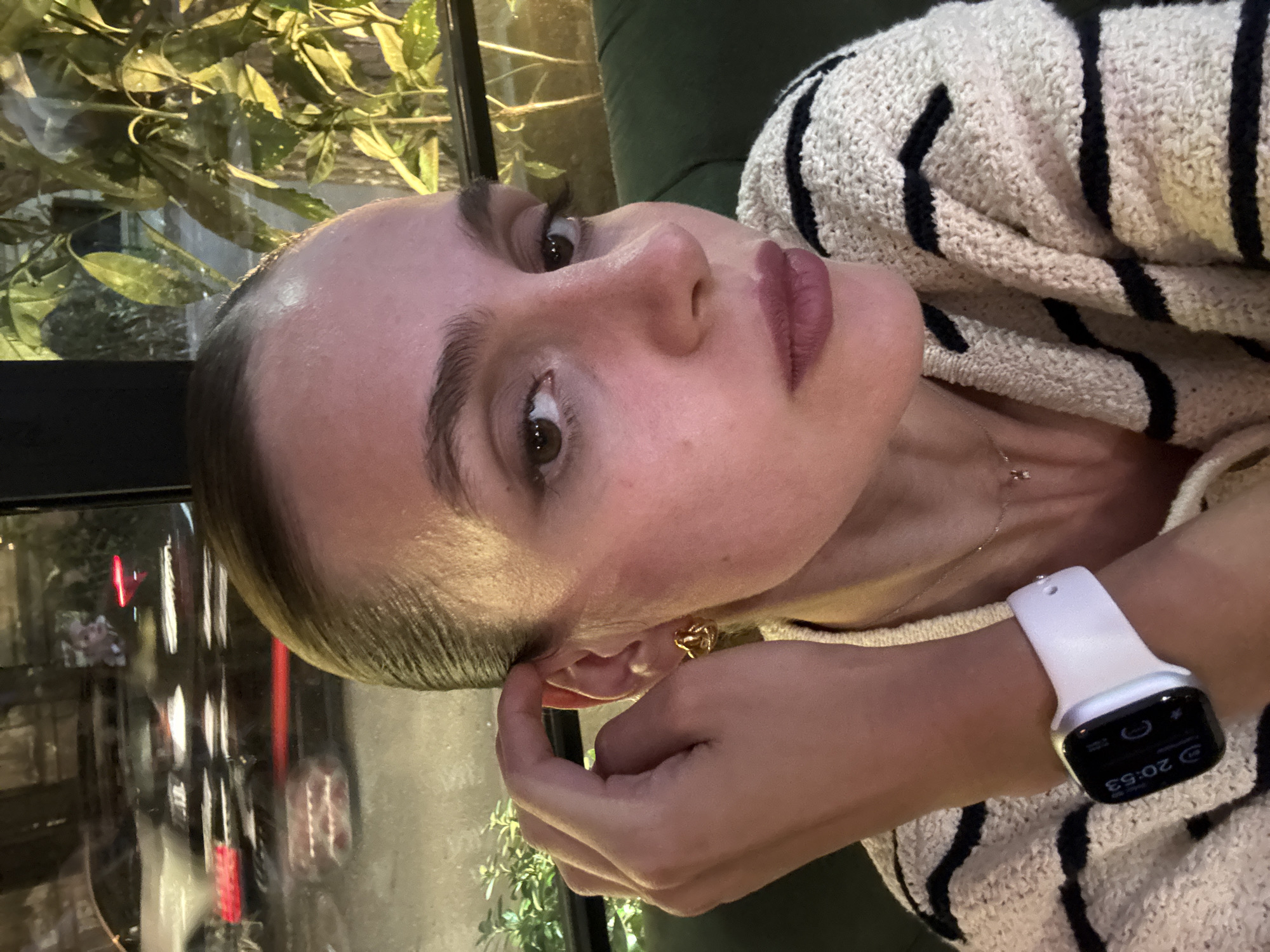 A close-up, eye-level portrait features a person with light skin, appearing to be female, looking directly at the viewer with a neutral, slightly serious expression. Their hair is slicked back, and they have defined eyebrows and what appears to be pinkish-mauve lipstick. The person is wearing a cream or off-white knitted sweater with bold black horizontal stripes. A delicate gold chain with a small charm is visible around their neck, and a small gold earring, possibly heart-shaped, is in their right ear.
Their right hand is casually placed near their right ear, while a white smartwatch with a dark screen is worn on their left wrist. The smartwatch clearly displays the time "20:53", indicating it is 8:53 PM.
The setting appears to be indoors, possibly a cafe or restaurant, at night. Behind the person, a dark green upholstered surface, likely a booth or chair, is visible. To the left, a large window reveals reflections of city lights and blurred red elements, suggesting a street scene outside. Green foliage or plants are visible through and behind the window, adding a natural element to the otherwise urban backdrop. The lighting is dim and ambient, highlighting the person's face while keeping the background softly lit. The overall atmosphere is intimate and quiet.