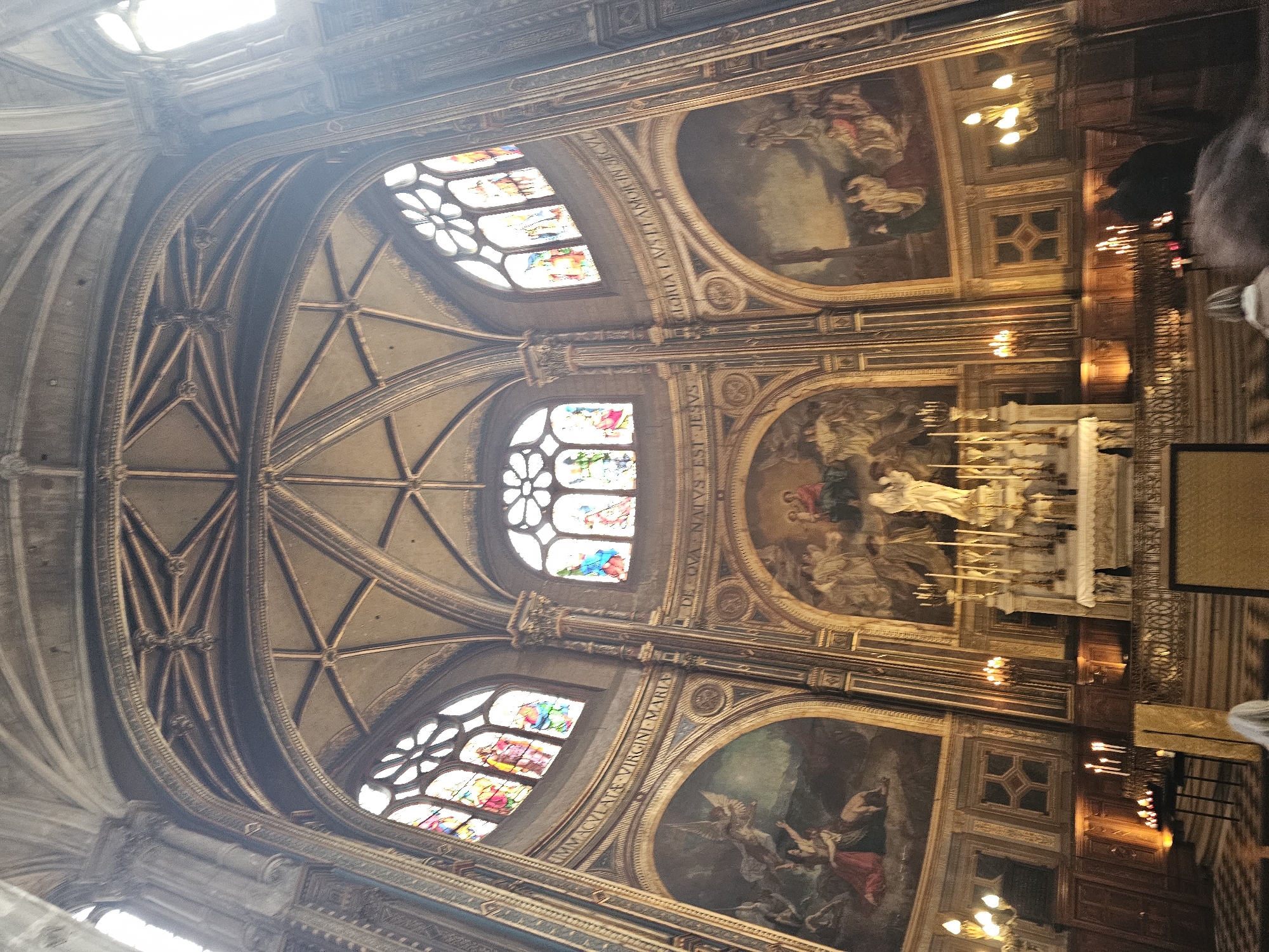 The image captures an interior view of a Parisian church, likely located in Paris, France. The camera angle is positioned looking upwards towards the ceiling, highlighting the architectural details and ornamentation. Stained glass windows provide colorful illumination. Paintings and artwork adorn the walls. The altar and candles add to the religious atmosphere. People are present in the scene.