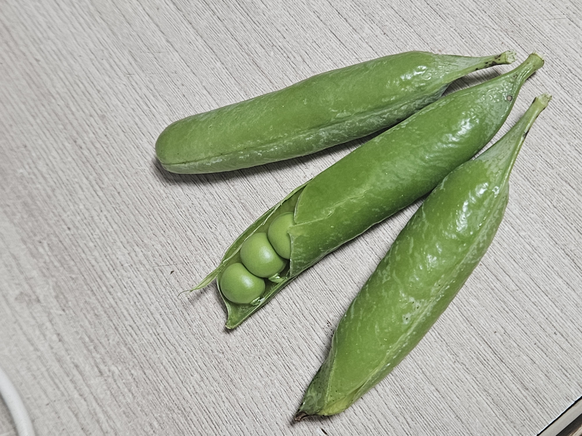 A close-up, overhead shot shows three green pea pods resting on a light-colored, wooden surface. One pod is open, revealing four round, green peas inside. The other two pods are closed. The surface has a visible wood grain texture. The lighting casts subtle shadows. The image contains no people or indication of location.