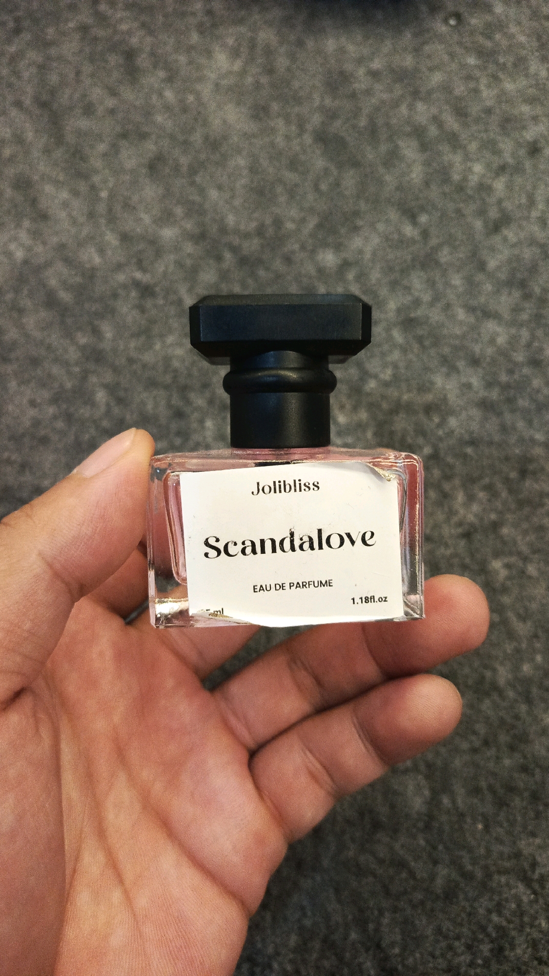 A close-up, overhead shot shows a person's left hand holding a small, clear glass perfume bottle with a black cap. The bottle contains a pink liquid. A white label on the bottle reads "Jolibliss" at the top, "Scandalove" in larger font, and "EAU DE PARFME" below, with "1.18fl.oz" to the right. The background is a textured gray surface. The image was taken on a gray, speckled surface.