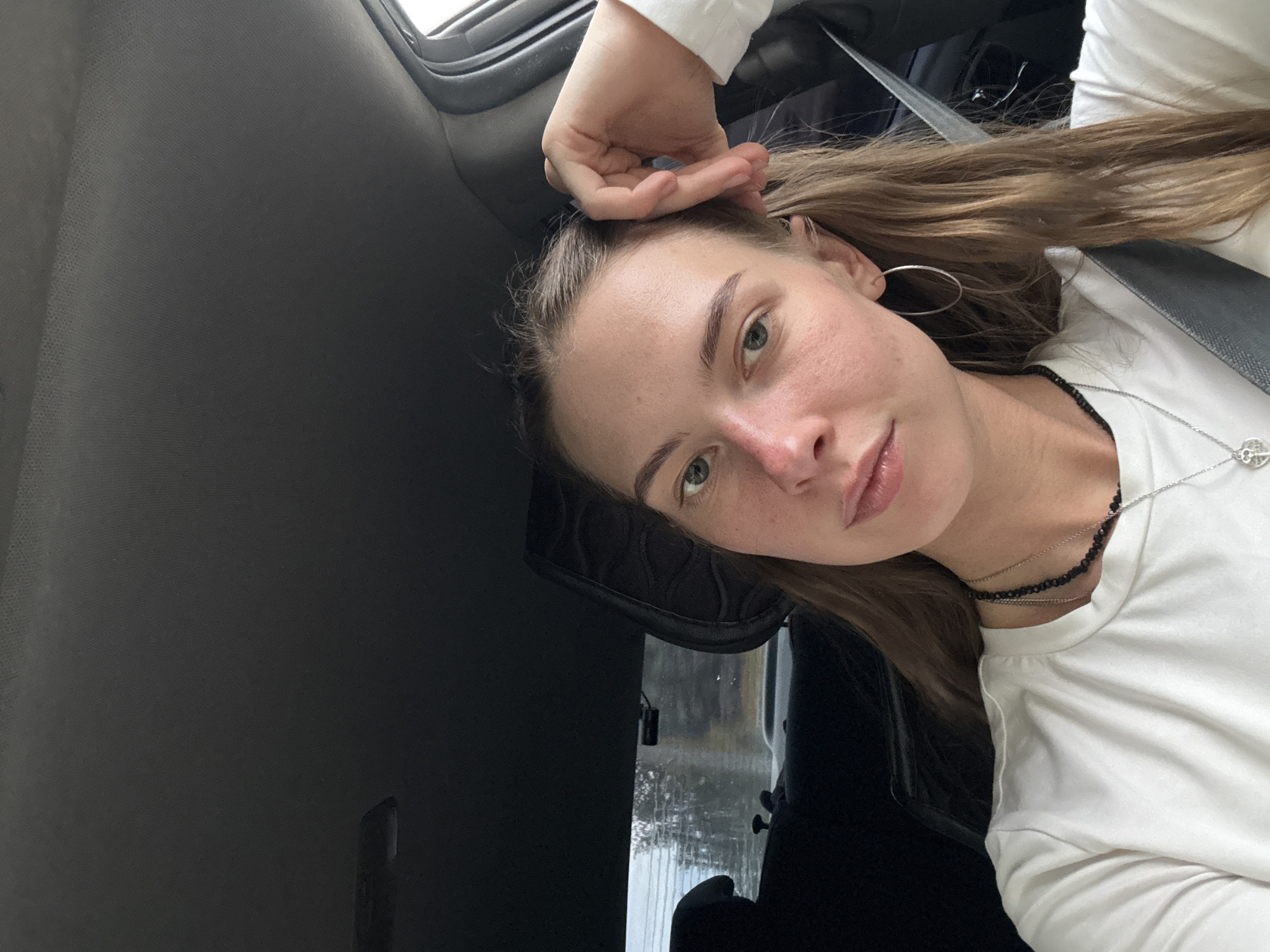 A young woman with fair skin and long, light brown hair is in the passenger seat of a car. She is looking at the camera, with her right hand resting on her head and her left hand slightly obscuring her face. She is wearing a white long-sleeved shirt, a black beaded necklace, and a silver pendant necklace. She also has a silver hoop earring in her left ear. The interior of the car is visible, with the headrest of the seat behind her and the car's ceiling. The background outside the car window appears to be trees and a cloudy sky. The location is not specified within the city of Perm, Russia.