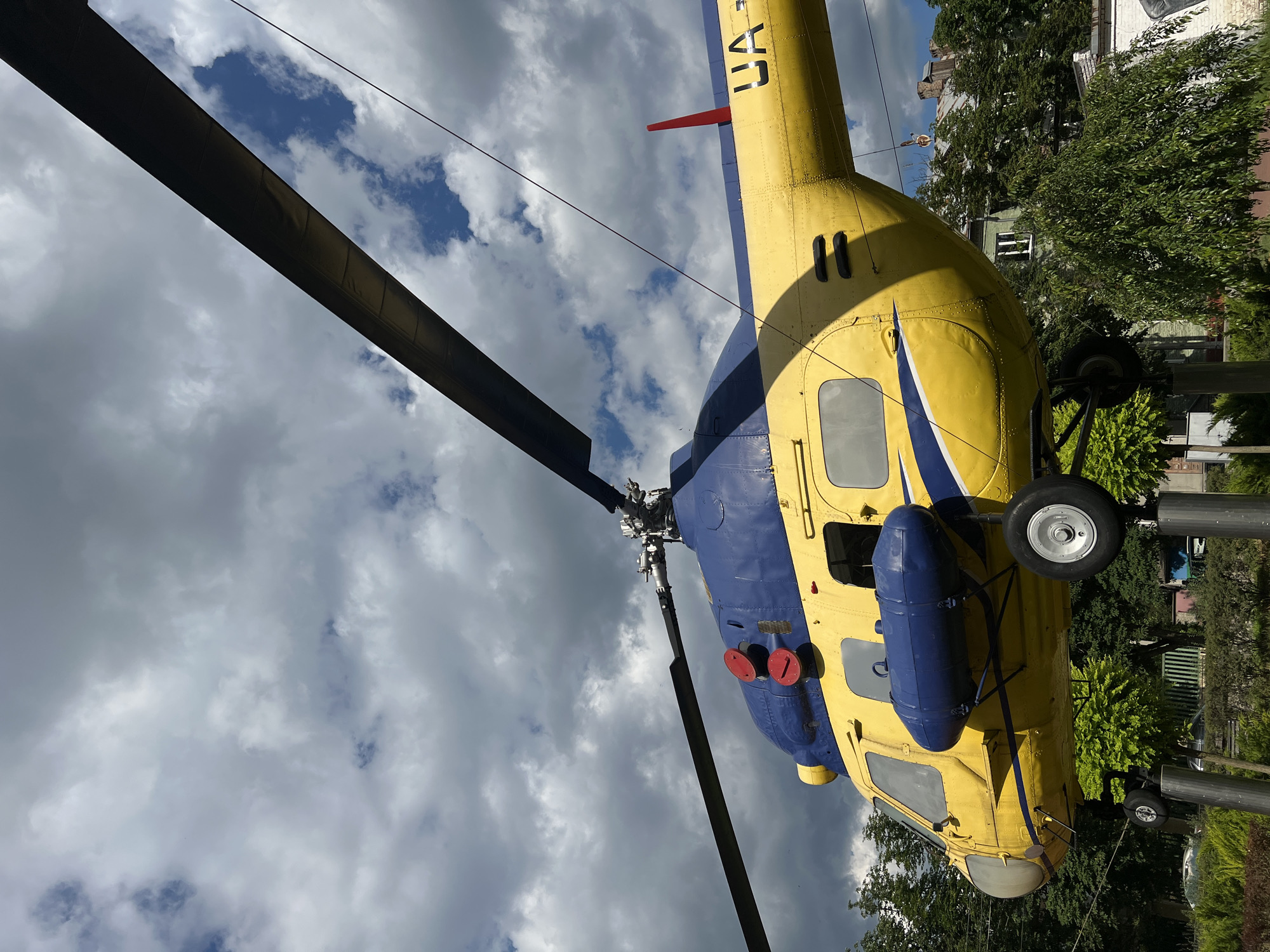 A yellow and blue helicopter is displayed on poles in an outdoor setting. The helicopter's rotor blades are extended, and the body is painted in bright yellow with blue accents. There are two red circular details on the top of the helicopter's body. A blue cylindrical tank is attached to the side of the helicopter. The helicopter is supported by two metal poles visible beneath its landing gear. In the background, there are green trees and buildings, with a cloudy sky overhead. The helicopter has "UA" printed on its tail.