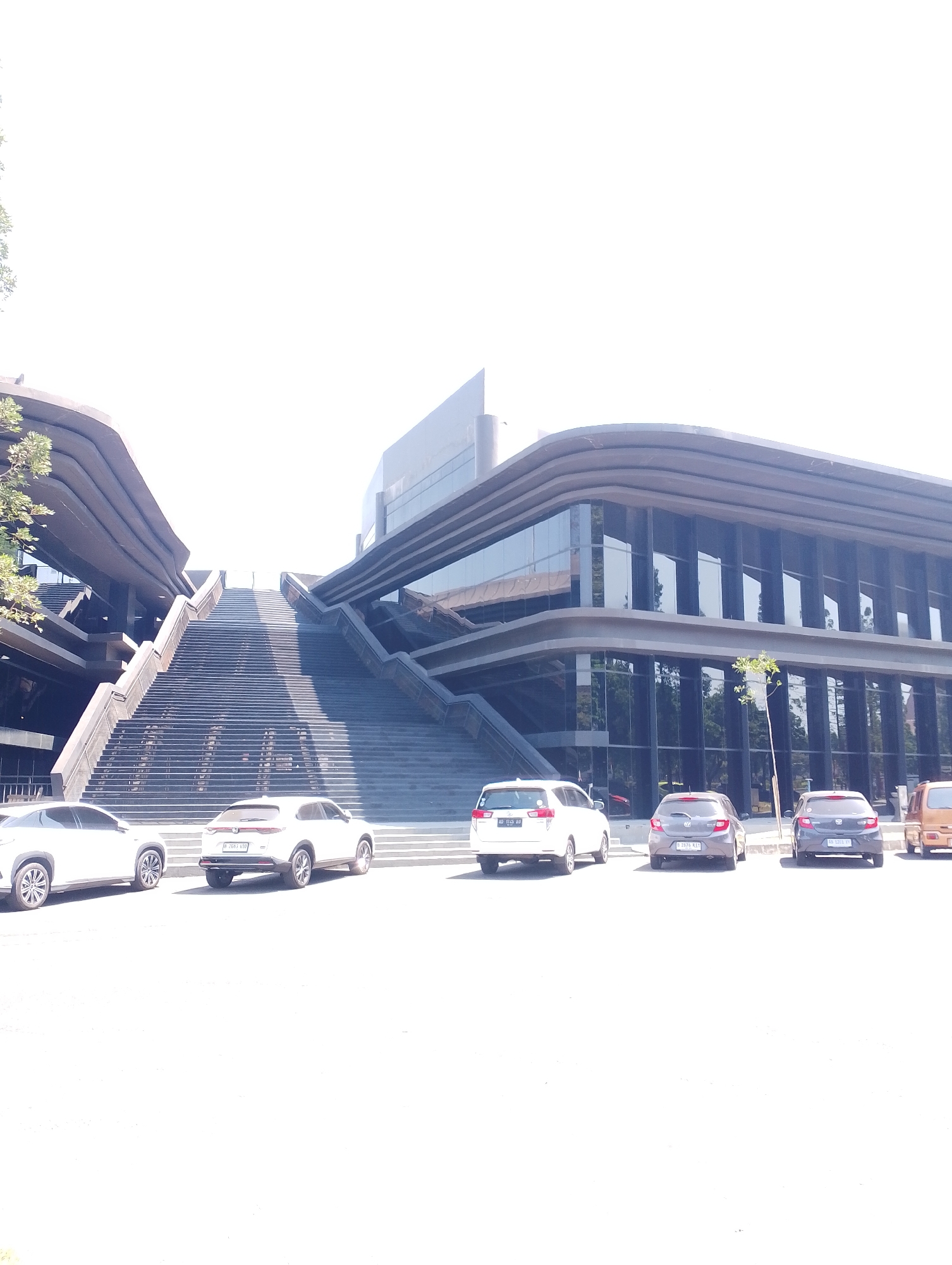 The image shows the exterior of a modern building with a large staircase leading up to it. Several cars are parked in front of the building. The building has a contemporary architectural style with a dark, curved facade and large glass windows. The sky is bright and overcast, suggesting daytime. The stairs are wide and appear to be made of dark stone or concrete, with many steps. The cars are of various makes and models, including a white SUV on the left, a white compact SUV in the middle, and a grey hatchback to its right. The location is identified as Depok, Indonesia.