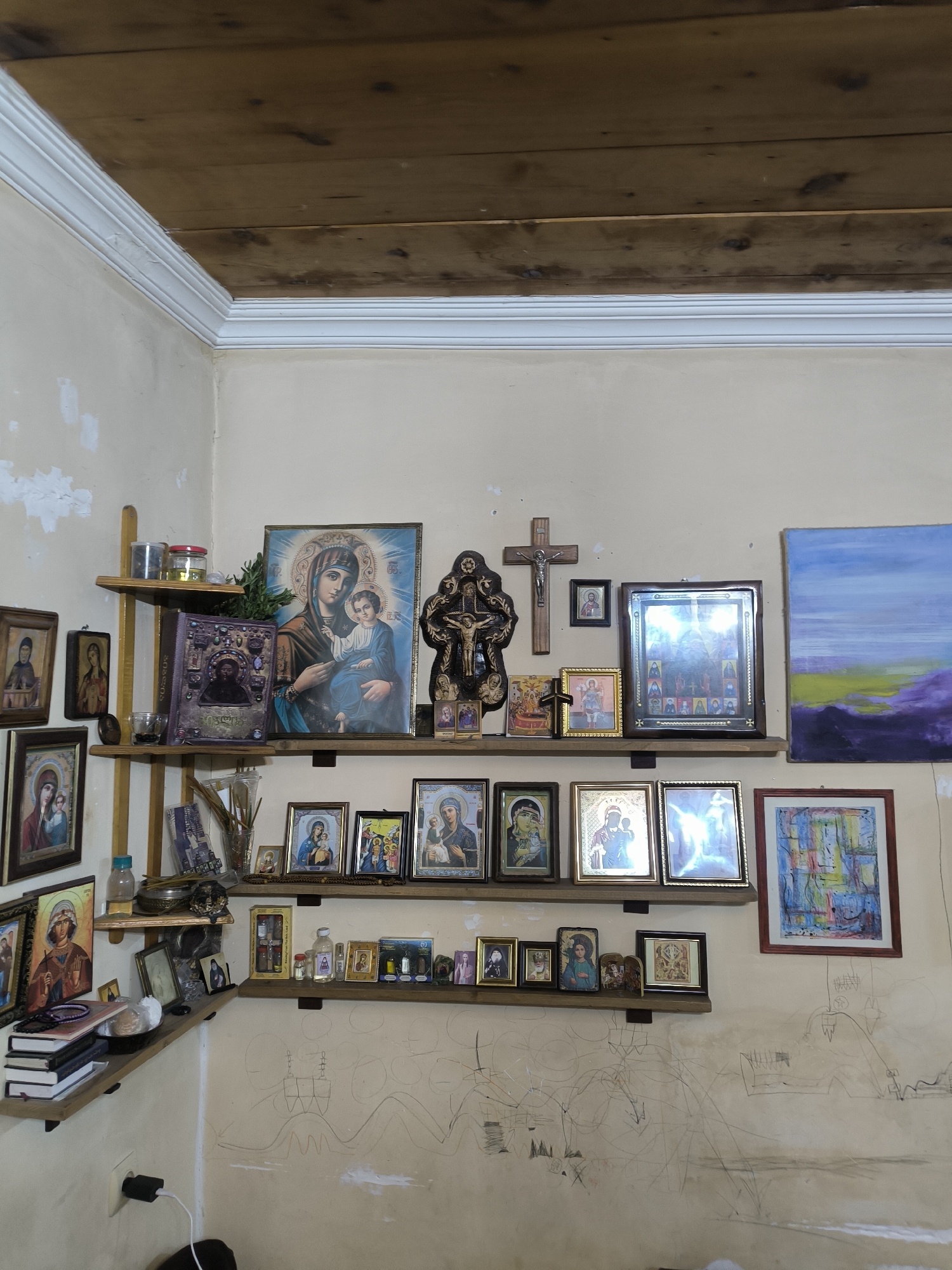 The image captures a corner of a room in Tbilisi, Georgia, characterized by an eclectic and spiritual atmosphere. Walls, painted in a faded beige hue, exhibit evidence of wear and tear, including patches of missing paint and seemingly random pencil markings.
A series of wooden shelves, attached to the walls, serve as platforms for an assortment of religious icons, framed images, and artifacts. The collection features depictions of the Virgin Mary and Jesus, as well as Orthodox crosses. Other objects like books, bottles, and smaller decorative items are scattered across the shelves.
The ceiling consists of exposed wooden planks, bordered by a decorative white molding. In the lower left corner, a power outlet with a charging adapter and a portion of what appears to be a black object (possibly clothing) are visible. The room is illuminated by an unseen light source, creating shadows and highlighting the texture of the surfaces.