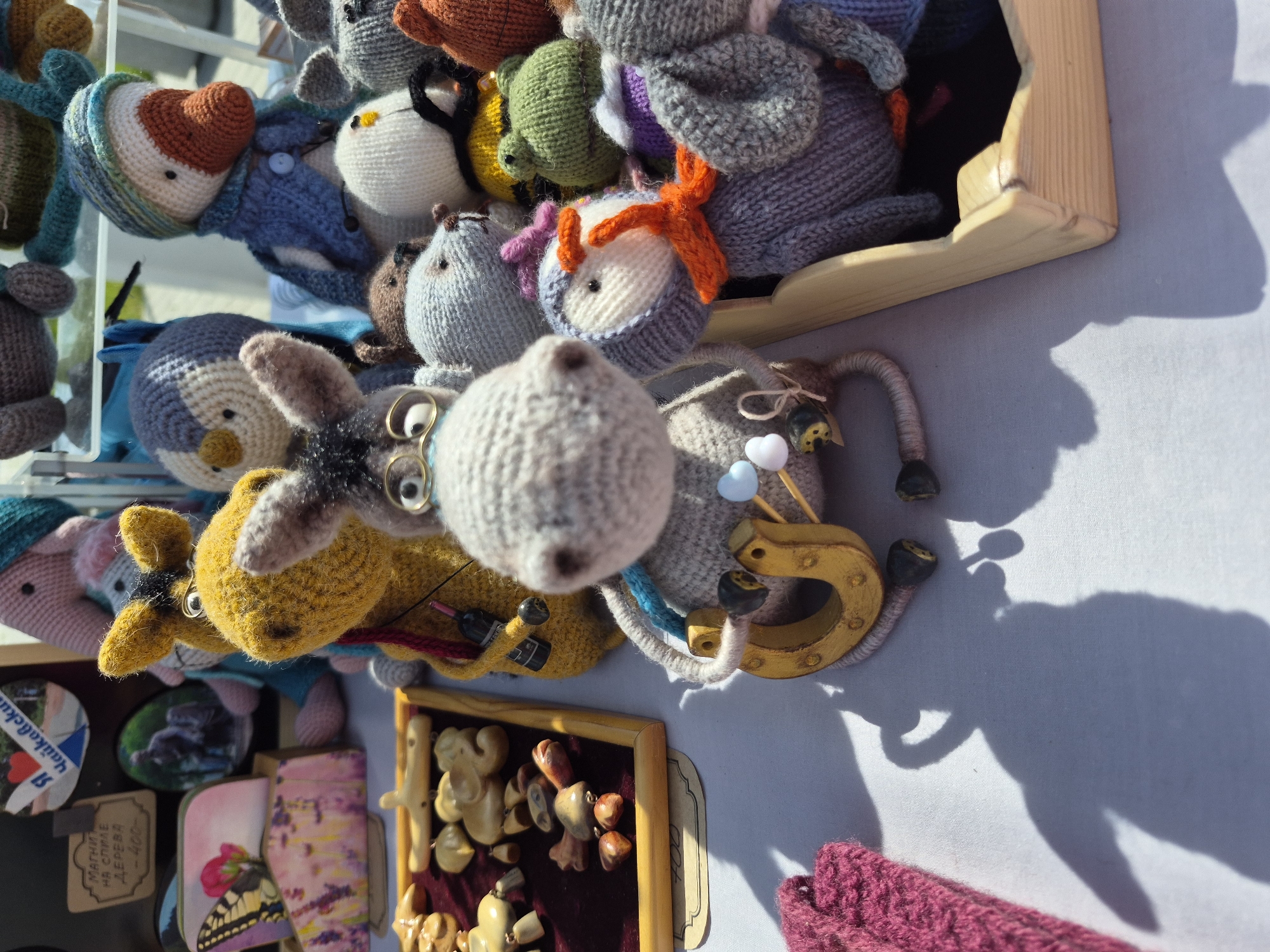 The image captures a close-up view of a collection of handmade, knitted animal toys displayed on a light-colored surface, likely a table or stall. The toys, which include various animals like mice, donkeys, and owls, are arranged in a slightly cluttered manner, suggesting a craft fair or market setting. A wooden box or frame contains some of the toys, adding a sense of containment and organization. Other objects like magnets and framed art or crafts are also visible, further indicating a marketplace environment. The bright sunlight casts shadows across the scene, highlighting the texture and detail of the knitted items. The exact location within the city of Unknown, Unknown cannot be determined from the visual information.
