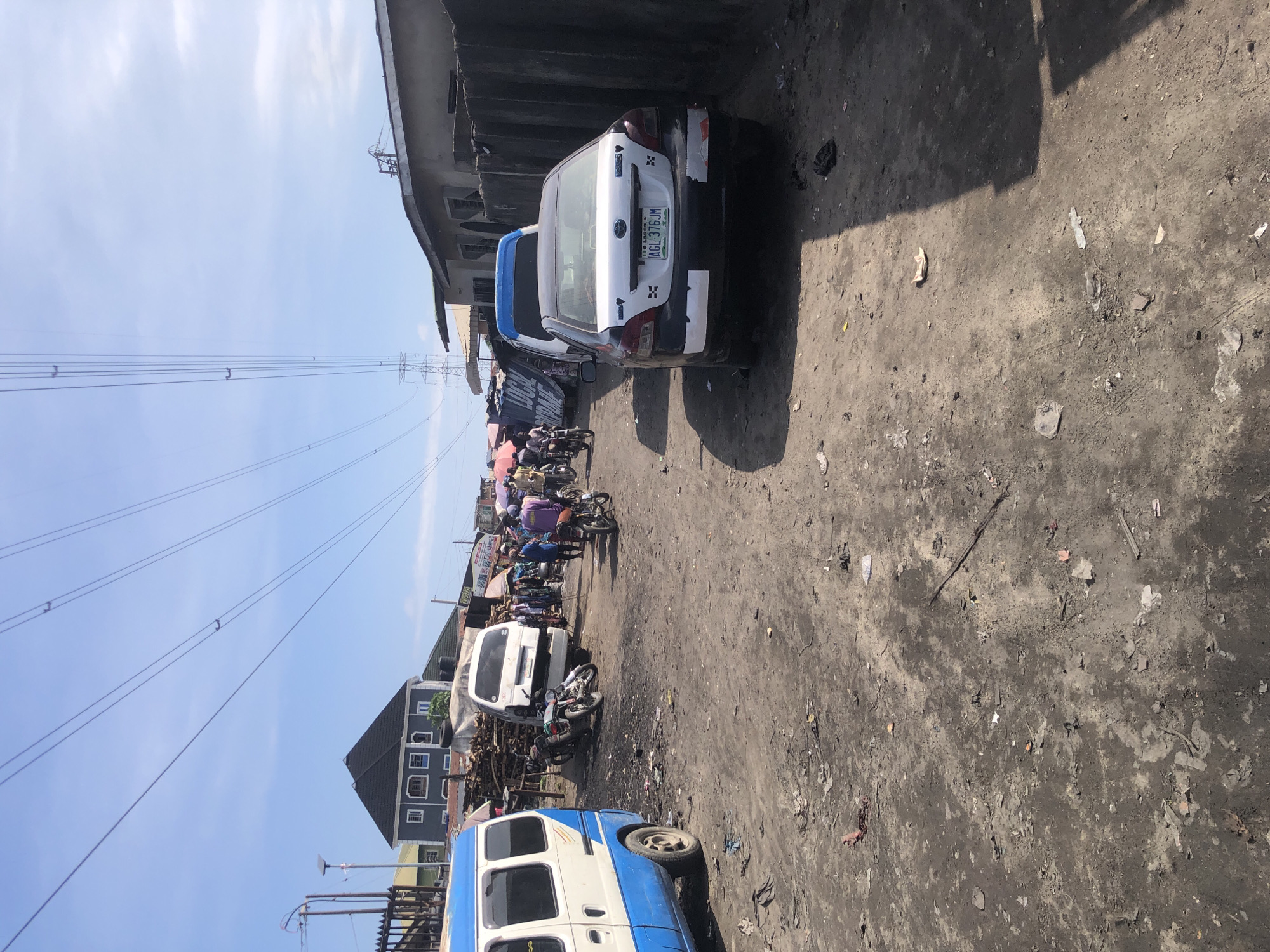 This is a daytime, eye-level, full shot photograph taken in a dusty, outdoor market area in Ado, Nigeria. The ground is covered in loose dirt, debris, and scattered litter. In the foreground, a white and blue van is parked on the left side of the frame. Further down the dirt road, several motorcycles are visible, some parked and others with riders. A white van is parked behind some stacked wooden materials.
To the right of the road, a dark-colored sedan with a license plate reading "AGL 376 JM" is parked. Beyond the vehicles, a line of market stalls with colorful awnings are set up. A number of people are walking and interacting within the market area, some appearing to be vendors and customers. The background features several buildings with corrugated metal roofs, and a prominent set of high-voltage power lines stretch across the sky, supported by transmission towers. The sky is clear blue with some wispy clouds. The overall scene depicts a bustling, informal marketplace in an urban or semi-urban environment.