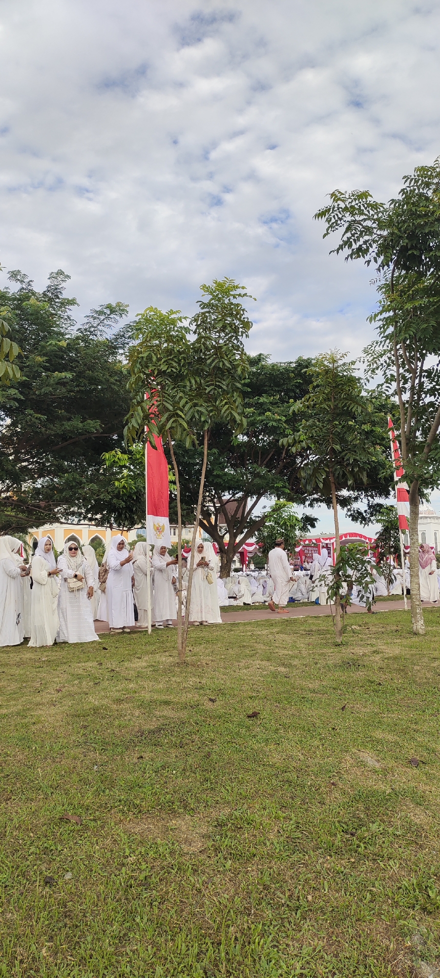 In the city of Meunasahaluedrienlhoksukon Barat, Indonesia, on a cloudy day with scattered white clouds, a group of people dressed in white attire are gathered in an outdoor area with a grassy foreground. The background features lush green trees and buildings with yellow roofs and white walls. Several red and white Indonesian flags are visible, one prominently displayed in the center of the frame. The individuals appear to be engaged in a religious or cultural event, with some standing in conversation and others seated on the ground. The scene suggests a communal gathering, possibly a religious ceremony or celebration, taking place in a park-like setting.
