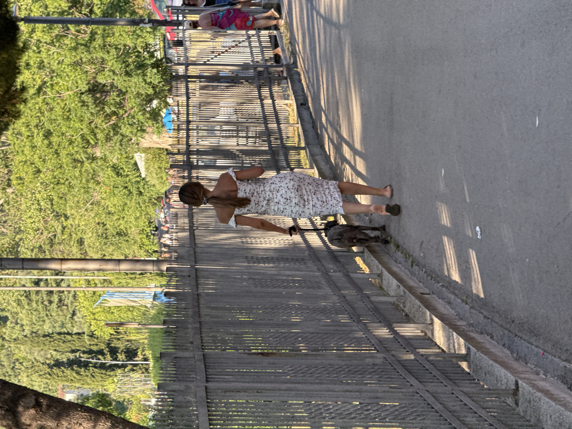 A woman in a floral print off-the-shoulder dress and flip-flops walks a grey pit bull dog on a leash down a paved walkway adjacent to a metal fence. Sunlight casts striped shadows across the walkway from the fence. In the background, beyond the fence, a crowd of people are visible, along with trees and a light pole. To the right, another woman in a colorful sarong is also walking, and a man with a white hat and sunglasses is walking behind her. The environment appears to be a public area, possibly a park or promenade, with lush greenery suggesting a warm climate. This scene could be the promenade along the coast in Budva, Montenegro, given the style of the walkway, fence, and surrounding vegetation.