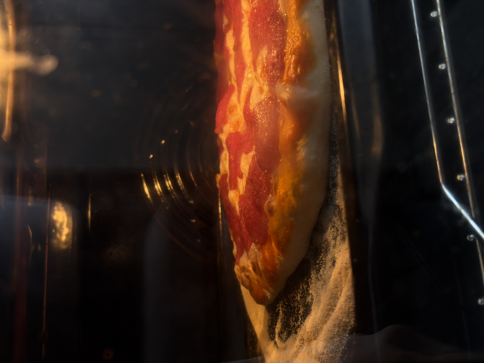 A pepperoni pizza is baking in an oven, viewed through the oven door. The interior of the oven is dark except where illuminated by the heating element. The pizza appears to be nearly finished baking. No people are visible. No city information can be derived from the image.