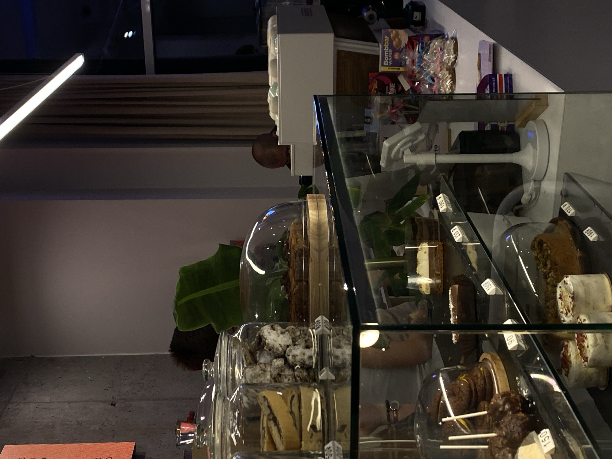 A dimly lit cafe in Vishnyaki, Russia, displays a variety of pastries in glass cases. In the foreground, a glass display counter holds several cakes and cookies. One cake, priced at 190 rubles, is topped with what appears to be nuts and a creamy frosting. Next to it, a round cake with red sprinkles is visible. To the left, a display case contains cookies and other baked goods. In the background, a person, possibly an employee, is seen from behind, with their head bowed, near a cash register. The cafe's decor includes concrete walls, large windows with beige curtains, and a bright overhead light fixture. Packaged snacks and treats are also visible on a shelf behind the display counter.