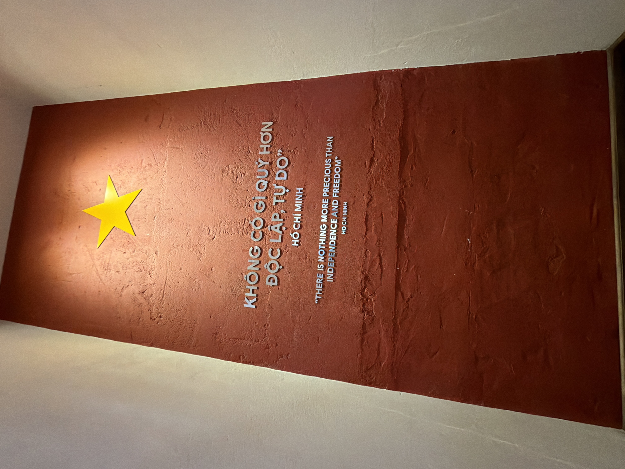 The image displays a textured, dark red wall with metallic lettering. At the top of the wall, a bright yellow star is affixed. Below the star, the text reads in Vietnamese: "KHÔNG GÌ QUÝ HƠN ĐỘC LẬP, TỰ DO” and then "HỒ CHÍ MINH". Below this, the English translation is given: "THERE IS NOTHING MORE PRECIOUS THAN INDEPENDENCE AND FREEDOM" followed by "HO CHI MINH". The wall appears to be a monument or display, likely commemorating Ho Chi Minh. The lighting suggests an indoor setting, possibly a museum or public space. The image does not provide specific information about the city of Da Nang, Vietnam, or any people present. The environment is characterized by the stark red wall contrasting with lighter-colored adjacent walls, forming a corner.