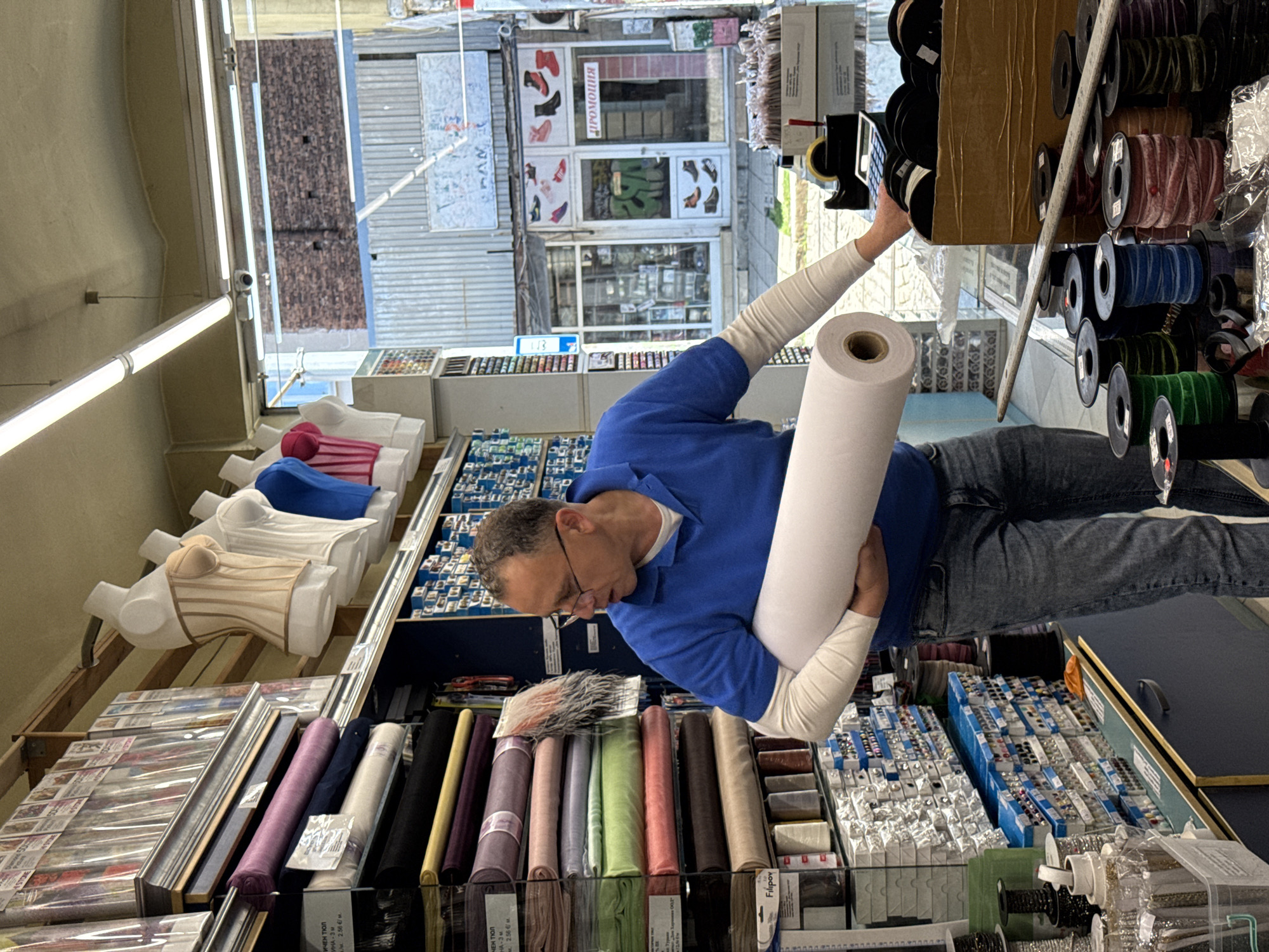 The photograph depicts a middle-aged man with short, gray hair and glasses standing in what appears to be a shop or store selling haberdashery. He is wearing a blue long-sleeved shirt and jeans. He is holding a large roll of white fabric, possibly cotton, and reaching towards a shelf filled with spools of ribbon of various colors and materials, some stored in a cardboard box.
The store is filled with an assortment of crafting and sewing supplies. There are mannequins displaying women's bodices, and shelves stacked with packaged fabric, ribbons, trims, and fasteners. A rack is visible, holding bolts of fabric in a range of colors.
Through the storefront windows, the exterior scene shows a building with signs advertising shoes, with text in Cyrillic, indicating the location might be in Bulgaria, most likely in the city of Sofia. Additional signage also indicates a "promo" or sales sign displayed in the window. The area outside appears to be a commercial district.