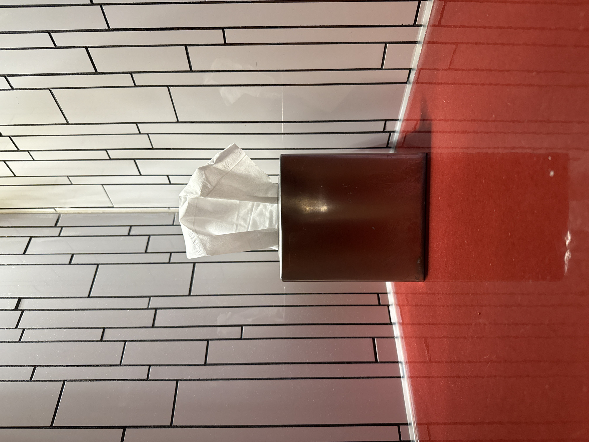The photograph shows a tissue box dispenser affixed to a tiled wall next to a red partition. The tiles are white with dark grout, laid in a horizontal, staggered pattern. The tissue box is dark brown and contains white tissues. The red partition is smooth and glossy, with horizontal lines that suggest a similar tile pattern, though the lines are more faint. The setting is in the city of Hollywood Seminole Reservation, United States of America.